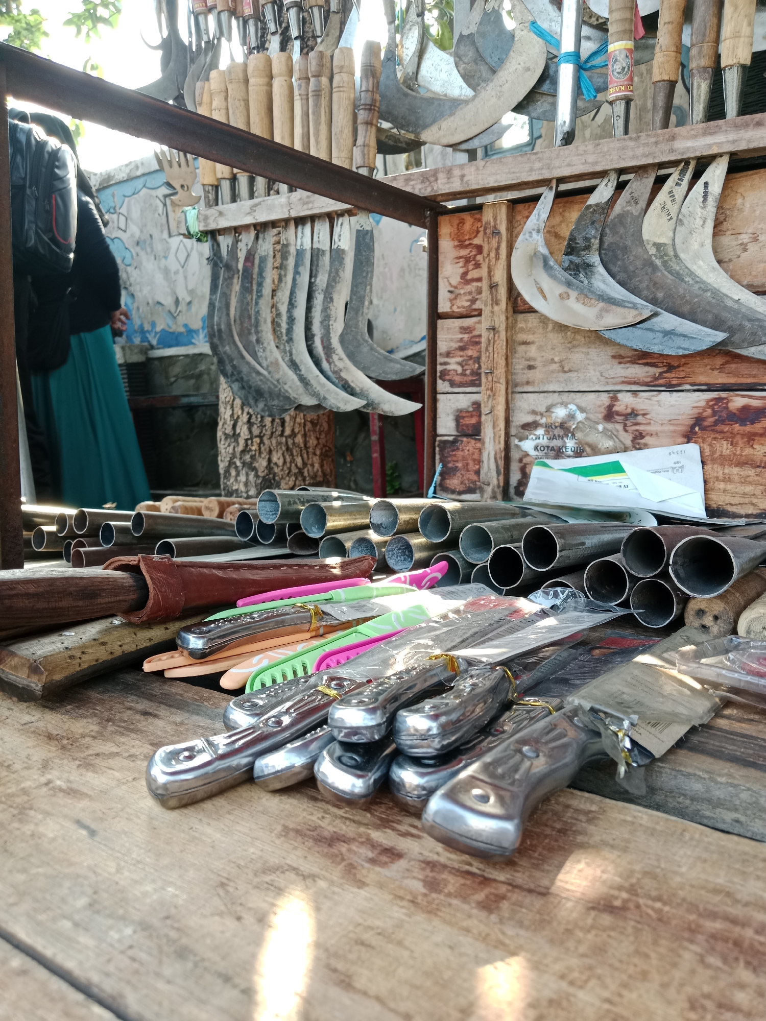 An outdoor stall displays an assortment of bladed tools and metal pipes. The stall appears to be set up on a wooden surface, likely a table, with a rustic wooden backdrop. In the upper portion of the image, a collection of curved bladed tools, possibly sickles or machetes, hang from a metal rack, some with wooden handles and others with metal. Below, a pile of metal pipes of varying sizes is stacked. In the foreground, several knives with metallic handles and colored plastic sheaths are arranged on the wooden surface. A brown leather sheath is also visible. In the background, a person wearing a dark top and a teal-colored long skirt is partially visible, along with a backpack. The wall behind the stall is decorated with a mural. A folded piece of paper, possibly an envelope, is placed on the wooden backdrop. The scene is lit by natural light, suggesting it is daytime. Based on the text visible on the envelope and backdrop, the location is identified as Kediri City, Indonesia.