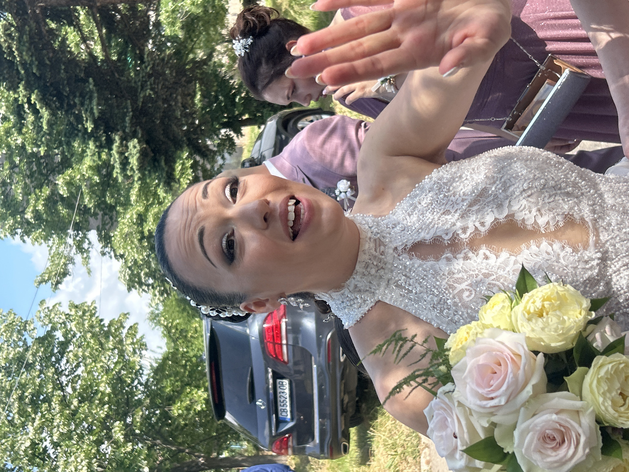 An outdoor, daytime scene in Buhovo, Bulgaria, features a woman in a wedding gown in the immediate foreground, with other individuals and a vehicle in the background.
The primary subject, a woman with dark hair styled in an updo adorned with pearl and crystal accessories, occupies the central and left portion of the image. She wears a white, halter-neck gown with intricate beadwork and lace detailing, featuring a deep V-shaped décolletage. Her facial expression shows her mouth open, revealing her teeth, with prominent white eyeliner on her upper lids. She wears dangling silver earrings with crystal elements. Her left hand is raised towards the viewer, palm open. In her left arm, she holds a bouquet of light pink and yellow roses, interspersed with green fern-like foliage.
To the main subject's immediate right and slightly behind her, a man is partially visible, wearing a light pink suit jacket with a white boutonnière. Further to the right and behind the main subject, a second woman is visible. She has dark hair in an updo with a white hair accessory and wears a purple or plum-colored dress. She is looking to her right, away from the primary subject, with a neutral expression. Her right hand holds a rectangular, metallic, box-style clutch purse, which is open and contains multiple banknotes. A white corsage is visible on her left wrist. A blue sleeve, possibly belonging to another person, is partially visible on the far left edge of the frame.
In the mid-ground, a dark grey Mercedes-Benz GLE SUV is parked, facing away from the viewer. Its rear license plate, "CB 5523 OB," is clearly visible.
The background consists of lush green trees with lighter green foliage, indicative of a warm season. A bright blue sky with scattered white clouds is visible above the tree line. The ground appears to be a mix of grass and dirt. Bright sunlight illuminates the scene, casting shadows on the subjects and objects.