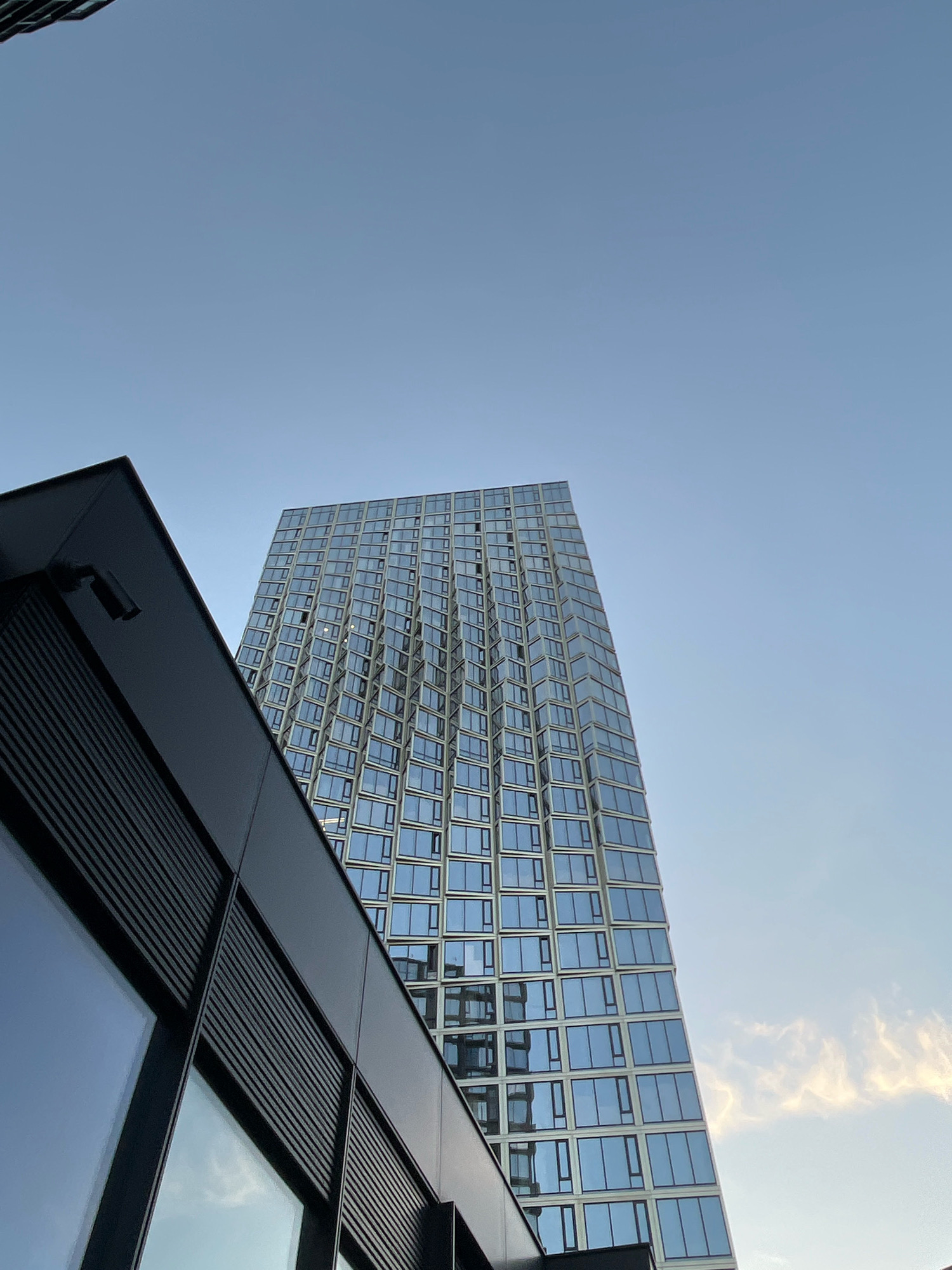 The image shows a low-angle view of modern buildings against a clear, pale blue sky with a hint of wispy clouds on the right. In the foreground, a dark, angular building with large reflective windows occupies the left side. Its facade features horizontal louvers or vents. Behind and to the right of this building, a tall, sleek skyscraper with a glass facade rises into the sky. The skyscraper's windows are arranged in a grid pattern, and the building appears to have a slight curve or wave-like texture to its exterior. The overall impression is one of contemporary urban architecture. The scene does not depict any people or specific activities.