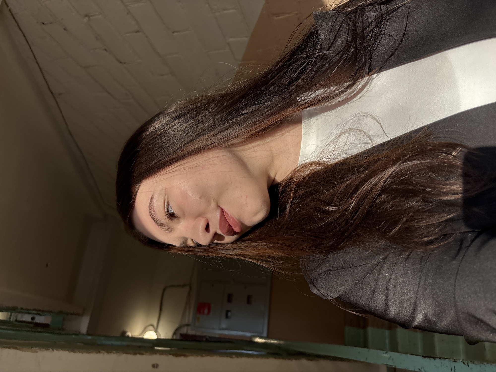 A woman with long brown hair is photographed indoors. She is wearing a white top under a black jacket. The location is indicated to be in Perm, Russia, but the specific location within the city is not evident from the image. There's a section of a painted brick wall visible, along with a set of pipes and a metallic electrical panel.