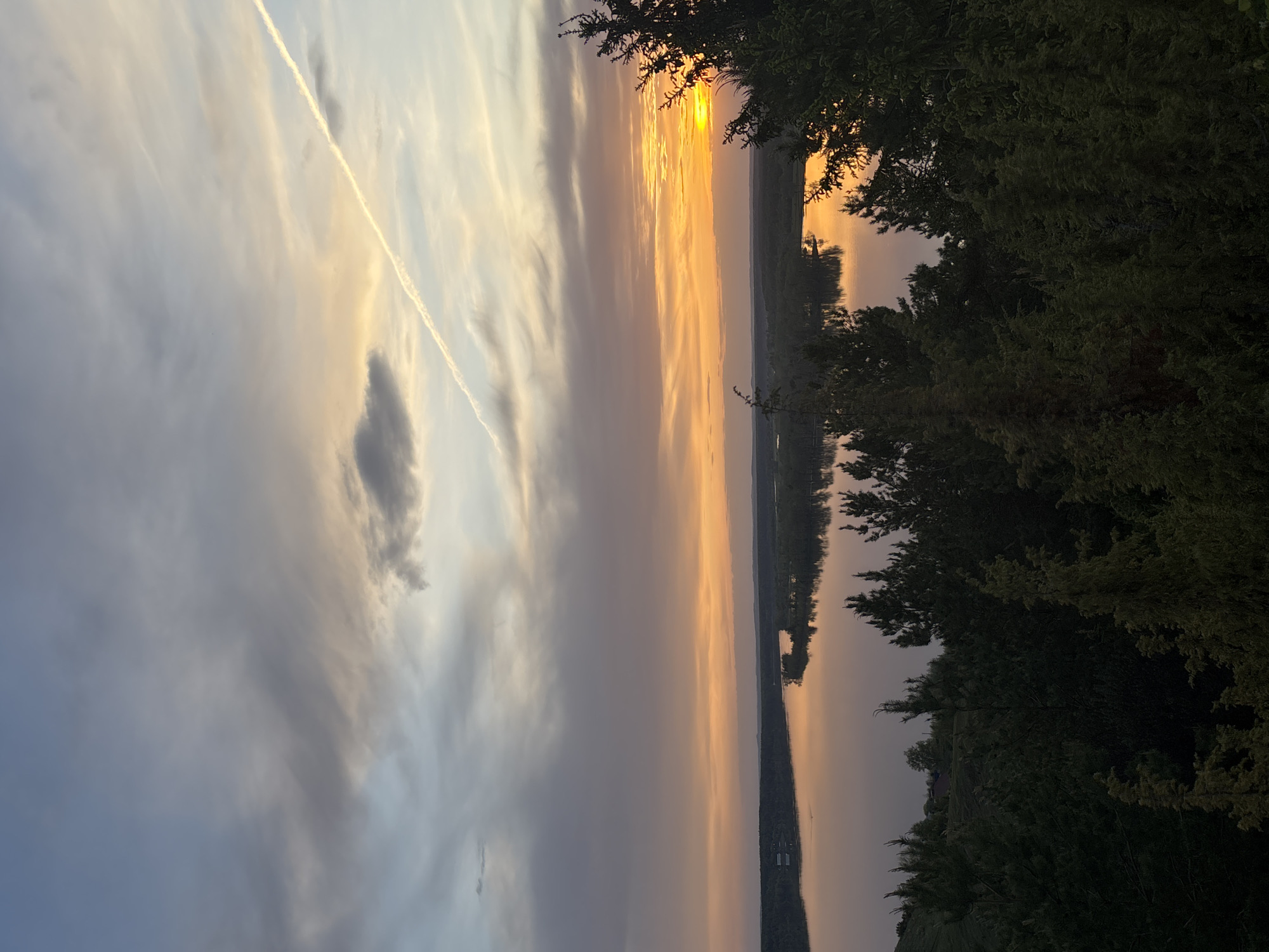 The image captures a sunset over a body of water, possibly a river or lake. The sky transitions from gray clouds on the left to a vibrant orange where the sun is setting on the right. A contrail streaks across the sky from the upper left. Trees are visible in the foreground on the right side of the image, silhouetted against the sunset. The water reflects the colors of the sky. There are no people or immediate objects visible. The location is suggested to be in Ольховка, Russia, but that cannot be confirmed with certainty from the image itself.