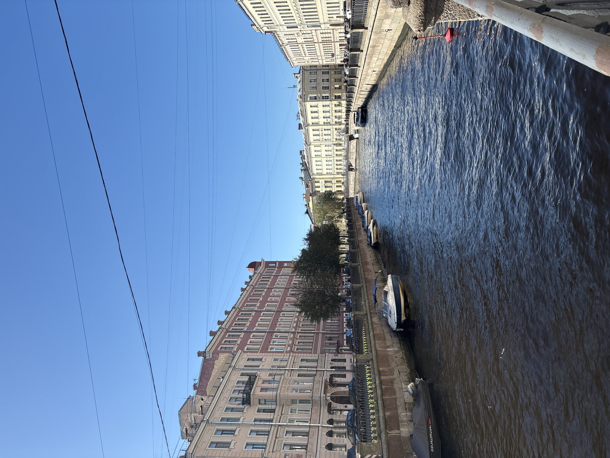The image shows a wide canal in Saint Petersburg, Russia, bordered by historic buildings. The buildings are multi-story, with ornate architecture and facades painted in various colors, including pink, red, and yellow. They line both sides of the canal, with windows, balconies, and rooftop details visible.
On the left bank, a black boat covered with a tarp is moored. Further along, a white and blue motorboat with its canopy up is tied to the canal wall. A black car is parked behind a decorative metal fence. Trees with dark green foliage are planted along the canal, providing a touch of nature between the urban structures.
On the right bank, the canal is also lined with stone walls and a decorative fence. Several boats are visible moored along this side as well, including a dark-colored boat further down. A red buoy with a black top floats in the canal. The water in the canal is a deep blue with visible ripples reflecting the clear, bright blue sky. Overhead, thin black wires are strung across the sky, indicating the presence of utility lines. The overall scene depicts a picturesque urban waterway, typical of Saint Petersburg's canal system, under a sunny sky.