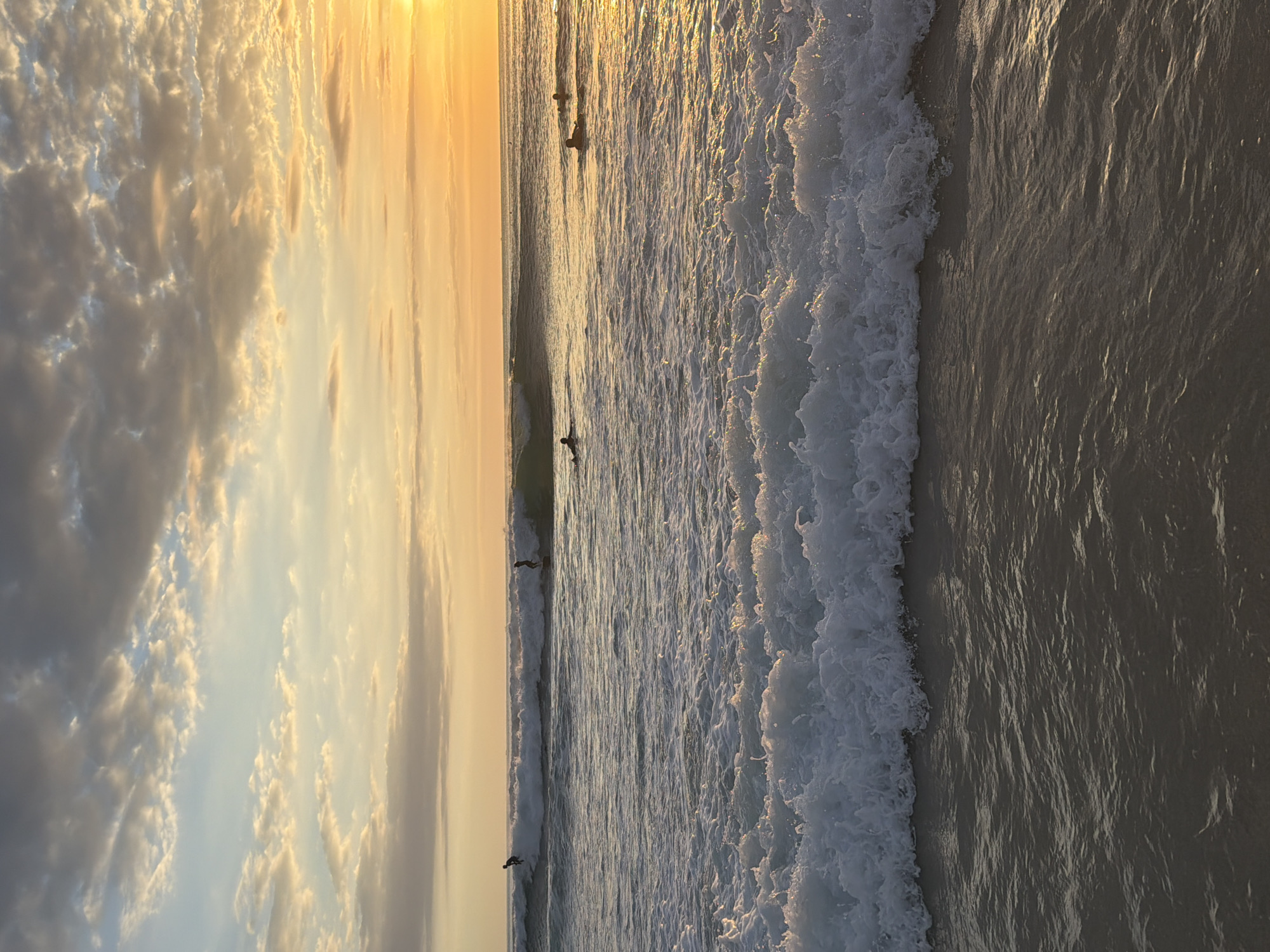 The image shows a beach in Kuta Selatan, Indonesia, at sunset. The sky is filled with golden and gray clouds. The ocean has waves breaking near the shore, and there are several individuals in the water, likely surfers. The sand is visible along the edge of the water.