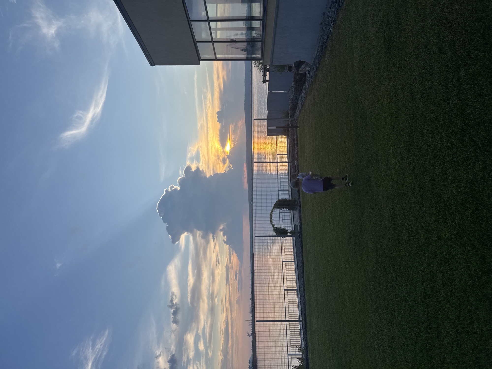 The image depicts a sunset scene on the Kama River in Chaikovsky, Russia. In the foreground, a well-maintained lawn area is visible, with several children playing. One child, wearing a purple t-shirt and dark shorts, is walking away from the camera. Further back, a modern building with large windows dominates the right side of the frame. A fence separates the lawn from the riverbank. The river itself is calm, reflecting the colors of the sunset. The sky is a dramatic display of blues and oranges, with distinct cloud formations, including a large, towering cloud with sunlight breaking through it. The overall atmosphere is serene and picturesque.