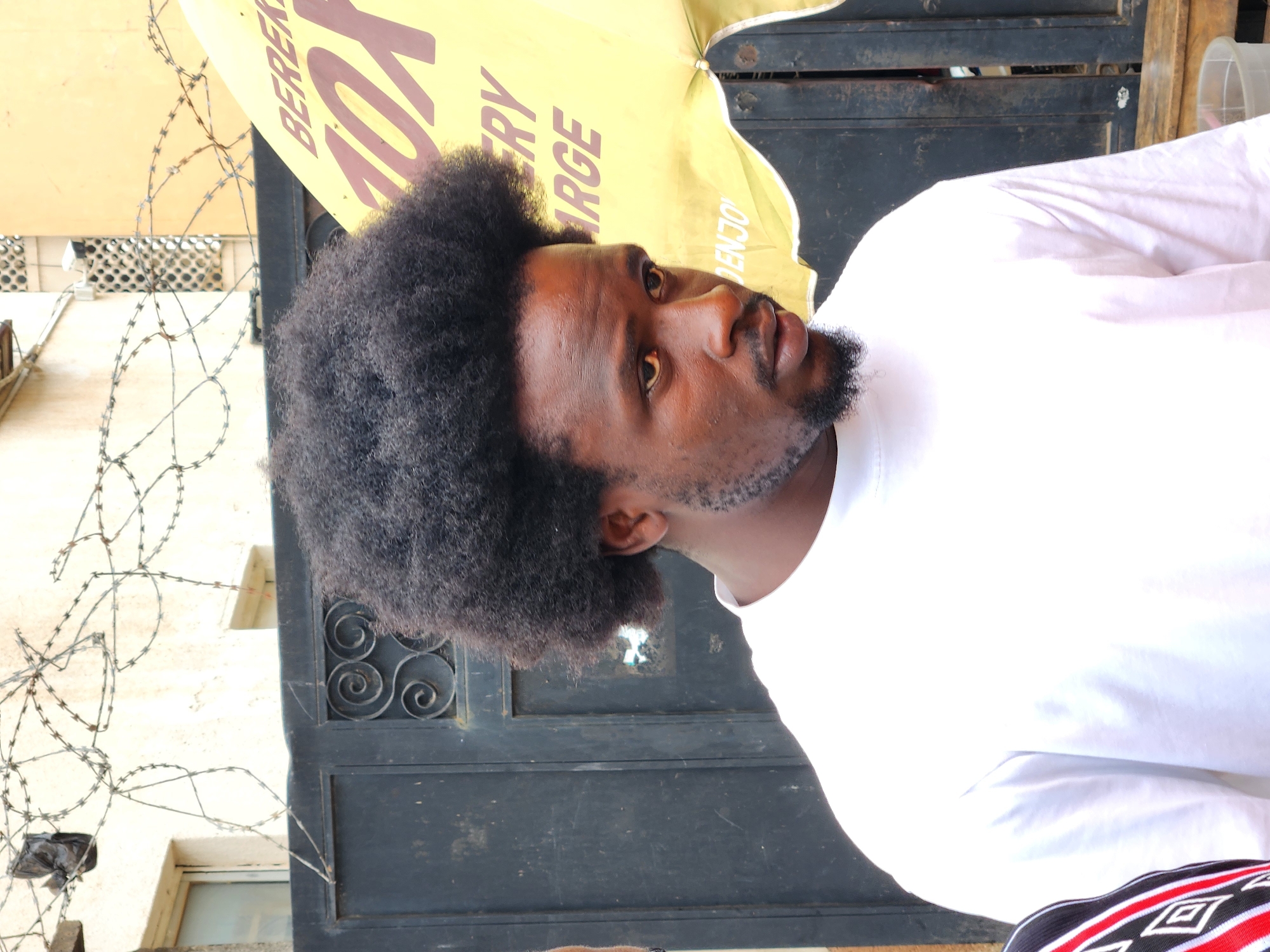 A portrait-style image, rotated 90 degrees counter-clockwise, depicts a man with dark skin, prominent dark curly hair in an afro style, and a trimmed beard. He is wearing a plain white t-shirt and looks slightly upwards and to his right with a neutral expression.
Behind and above the man's head is a bright yellow umbrella or canopy, partially visible. Text printed on the canopy includes "BERERE," "10X," "VERY," "LARGE," and "ENJOY," suggesting a promotional item for a product or business, possibly food-related given "BERERE" which is a known rice brand in Nigeria.
In the background, a dark, heavy metal gate or door with decorative scrollwork is visible, positioned next to the man's left side (viewer's left). Barbed wire is strung in complex patterns across the left side of the frame, indicating a security feature. Behind the barbed wire and gate, a light-colored building or wall with small, barred windows is discernible.
The lighting is bright and natural, suggesting it is daytime and sunny. A small section of patterned fabric, red, black, and white, is visible at the very bottom right of the image, possibly belonging to another person or object out of frame. The scene appears to be an urban or market setting, consistent with the provided context of Jos, Nigeria.