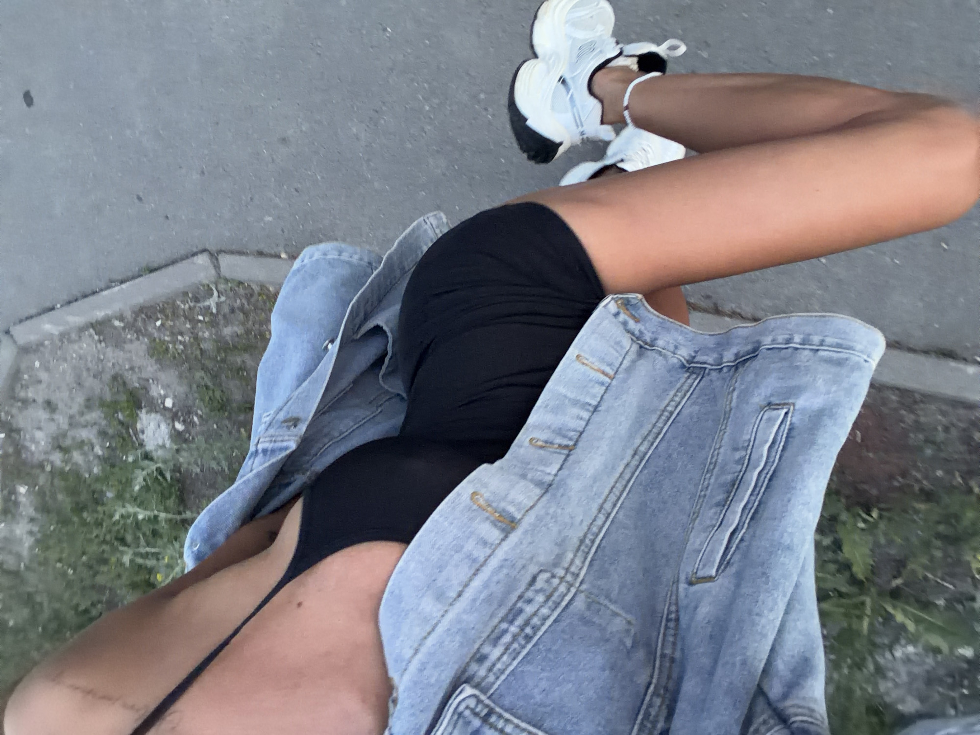 The image shows a close-up, high-angle shot of a person wearing a black tank top, a denim jacket, and white sneakers. The person is standing or walking on a paved surface, likely a sidewalk, next to a grassy area. The lighting suggests it is daytime, possibly late afternoon or early evening. No identifiable landmarks or signs are visible to pinpoint the location within Kazan, Russia. The focus is on the person's clothing and legs, with the background blurred.