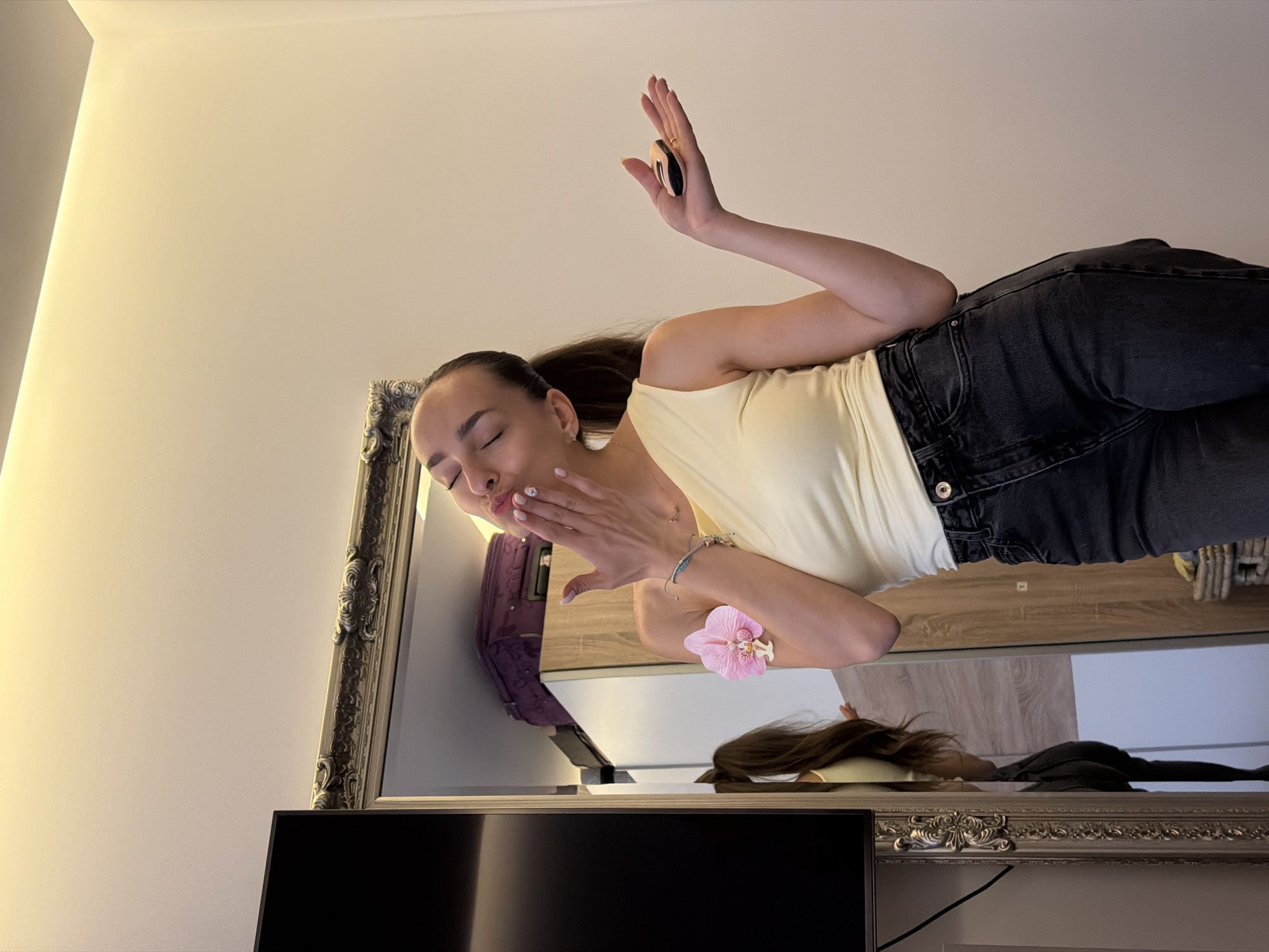 The image shows a young woman with her eyes closed, blowing a kiss, with a pink orchid pinned to her arm. She is wearing a light yellow one-shoulder top and dark wash denim jeans. In her right hand, she is holding a small, round cosmetic compact. She appears to be standing in front of a large, ornate mirror, which reflects her image and part of the room behind her. The reflection shows a purple suitcase on a shelf and a black television mounted on the wall. The room has a plain, light-colored wall and a ceiling with lighting fixtures. The overall impression is that of a personal dressing area or bedroom. There is no visual information in the image to suggest this location is in Sofia, Bulgaria.