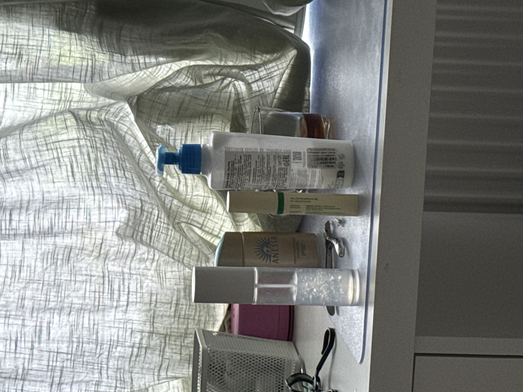 The image shows a desk with several cosmetic products and accessories, placed in front of a light-colored curtain. From left to right, there is a white mesh basket, a pink rectangular box, a clear spray bottle with a white cap, a cylindrical bottle with a tan label that reads "ANESSA", a small tube with green and white labeling, and a clear glass with a dark liquid. A pair of black-handled tweezers and a silver nail clipper are also visible on the surface. The lighting suggests it is daytime, with sunlight filtering through the curtain. The desk appears to be a vanity or dressing table. The location of Коробицыно, Russia, is not discernible from the image content.
