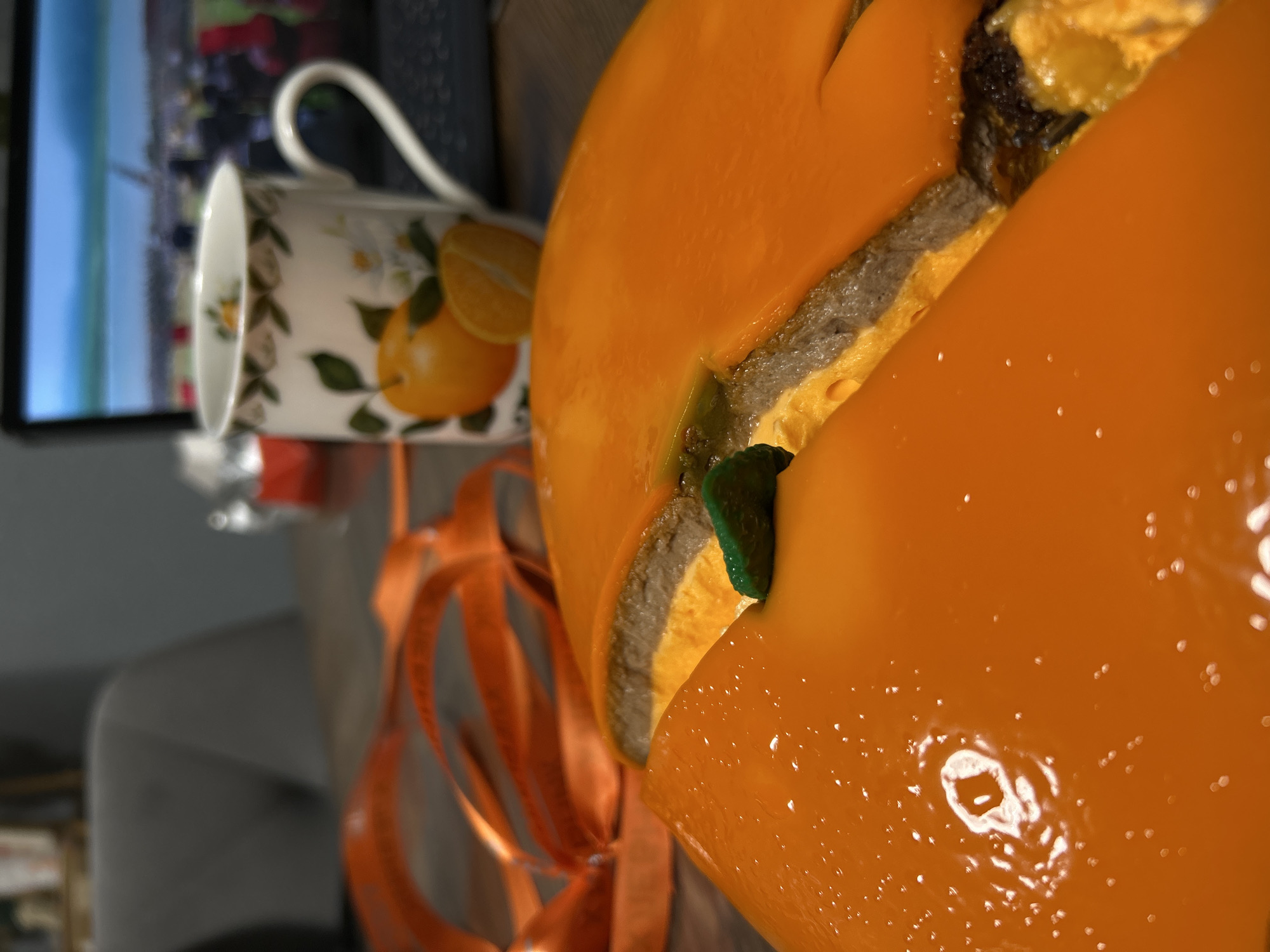 The image shows a still life with a large orange cake, sliced to reveal layers of sponge cake and filling. Next to the cake is a teacup adorned with lemon motifs and an orange ribbon. In the background, a television screen displays an out-of-focus scene that seems to show an urban shoreline with colorful structures. The location cannot be definitively pinpointed as Saint Petersburg, Russia, without additional context or details to confirm landmarks or geographical features specific to that city.