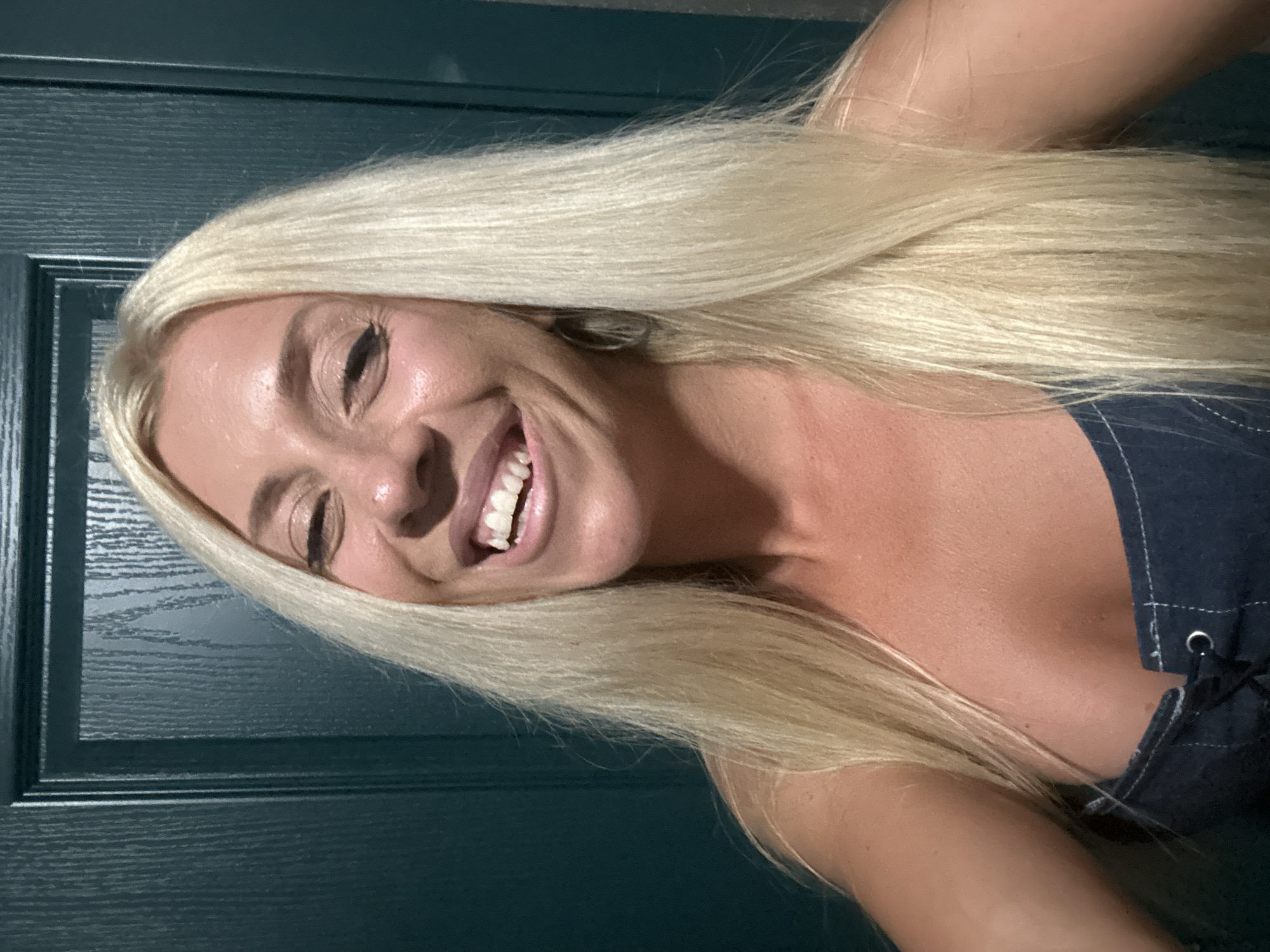 A woman with long blonde hair and a denim top is smiling at the camera in front of a dark green door. She is wearing makeup, including eyeliner and lipstick. The door has a vertical wood grain texture and a rectangular panel inset. The lighting is bright, highlighting the woman's skin and hair. The image was taken in Pazardzhik, Bulgaria.