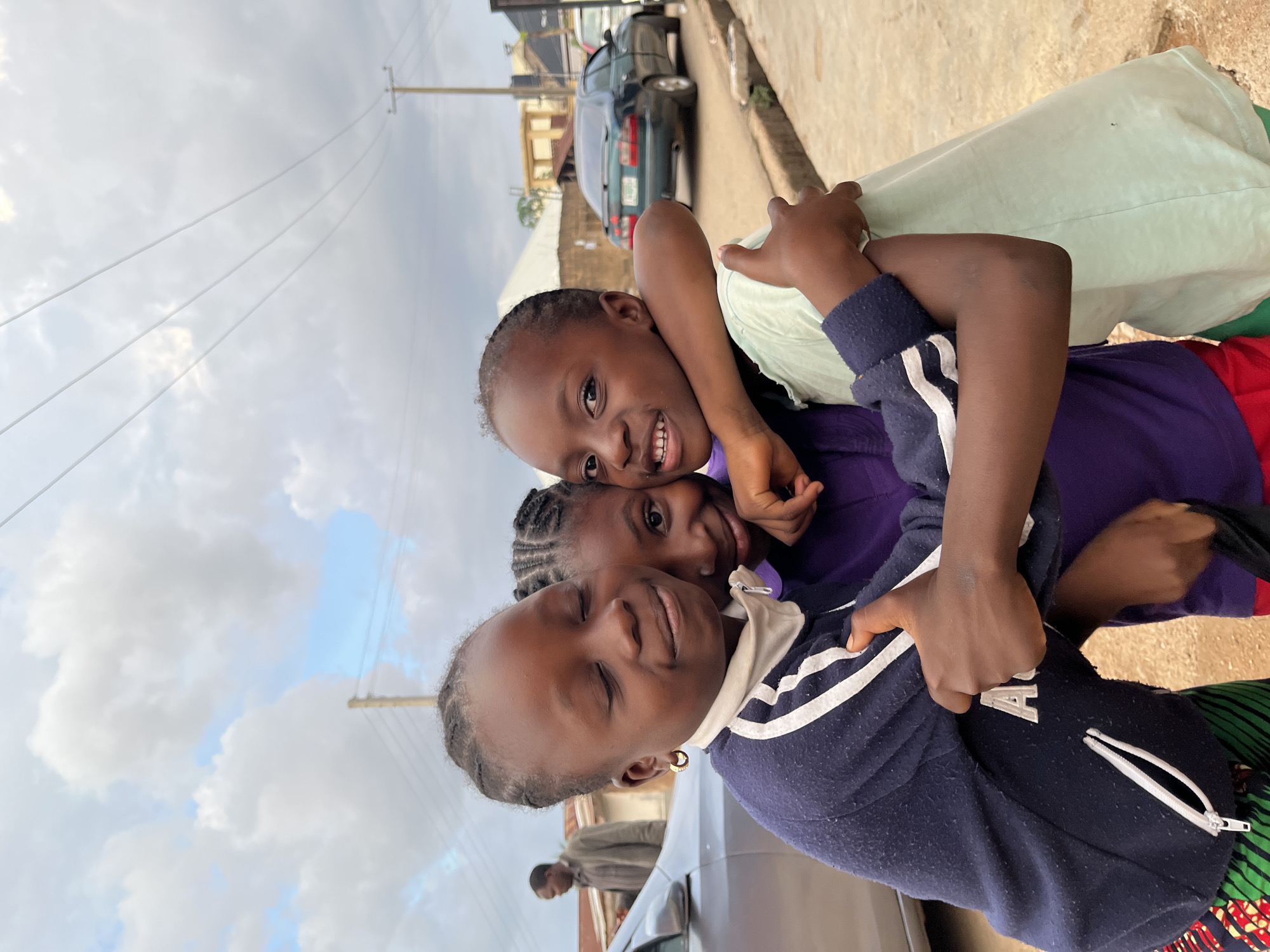 Three young girls are captured in a cheerful outdoor portrait, embracing each other on what appears to be a street in Jos, Nigeria. The girls are smiling broadly and looking directly at the camera. The girl in the foreground on the left wears a navy blue top with white stripes and a light collar, while the girl on the right is in a light green top over a purple garment. Another girl is nestled between them, partially visible. Their expressions convey joy and affection.
The scene is set on a paved street, likely a residential or urban area. In the background, a dark-colored sedan is parked along the roadside, and parts of low-rise buildings are visible. A utility pole and several power lines stretch across the upper part of the frame against a partly cloudy sky, indicating it is daytime. A man is faintly visible in the distance, partially obscured by a vehicle. No discernible text is clearly legible on any of the objects or clothing.