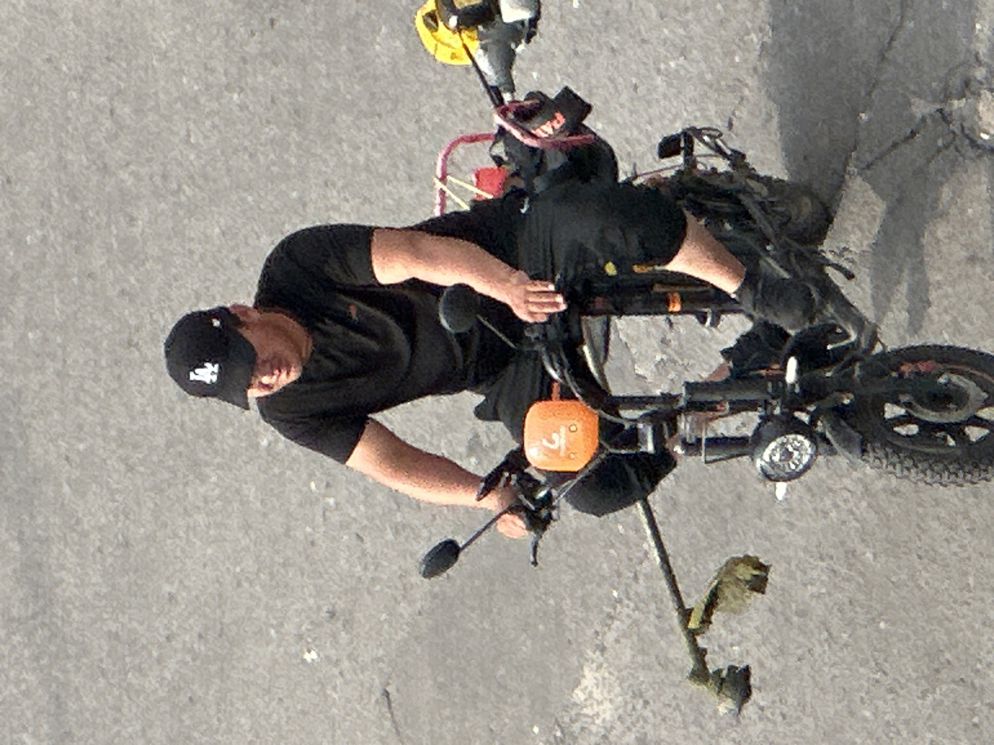 A man wearing a black baseball cap and a black t-shirt is riding an electric scooter on a paved surface, possibly a street or sidewalk in Podolsk, Russia. The man is seated on the scooter, with his legs positioned in front of him. He is holding onto the handlebars, and his right hand is near the throttle. Attached to the front of the scooter, to the left, is a string trimmer, with its cutting head extending outwards. To the right of the man, a yellow object, possibly part of another tool or equipment, is mounted on the scooter. A red plastic container is also visible behind the handlebars. The scooter has a headlight and a front wheel with a tread pattern. The background is a textured gray surface, indicative of pavement.