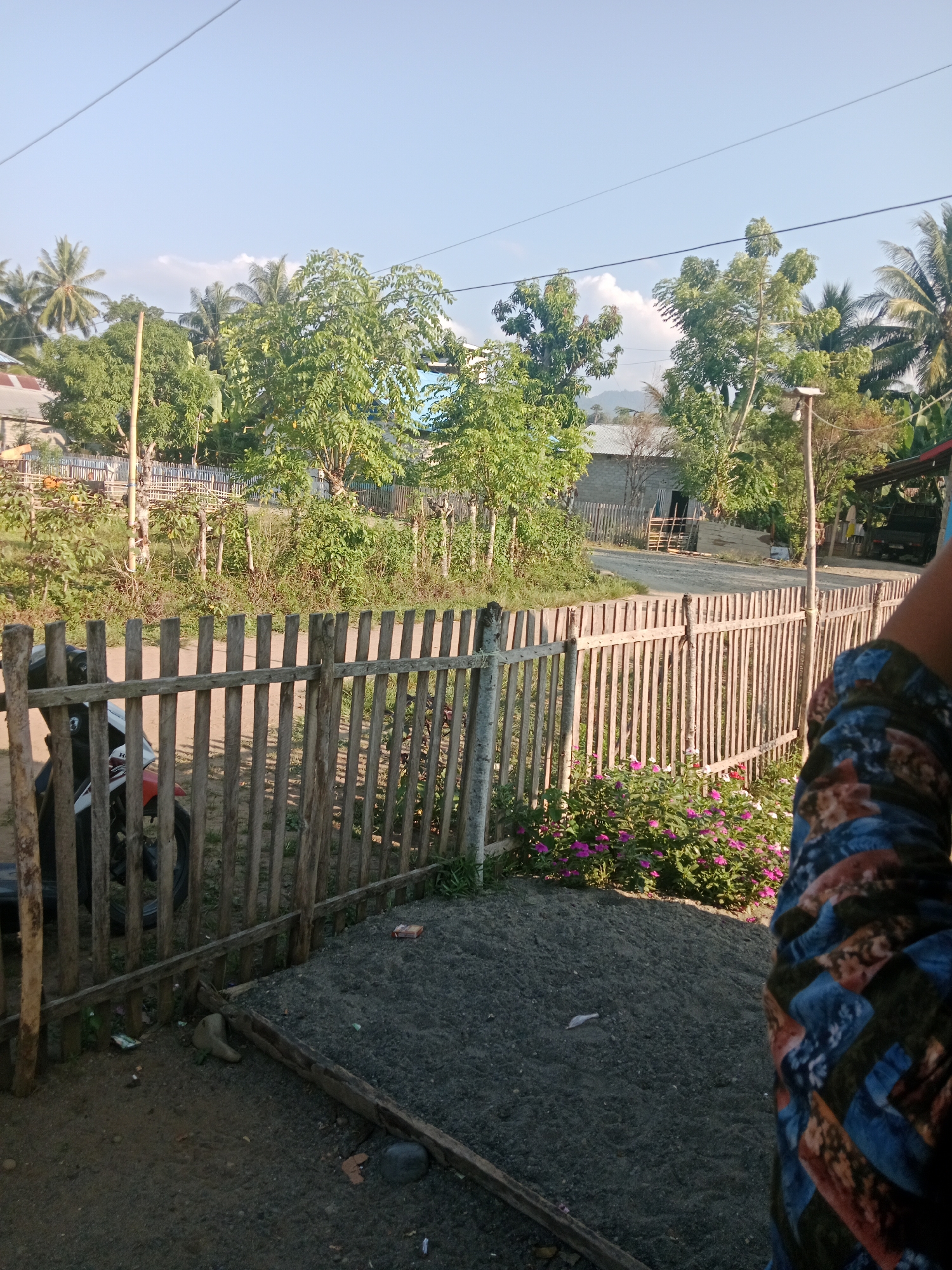 The image depicts a rural scene in Lakea Satu, Indonesia, under a clear blue sky with scattered clouds. A wooden fence made of vertical posts and horizontal planks runs through the foreground and middle ground, creating a barrier. To the left of the fence, a portion of a dark-colored motorcycle is visible, suggesting it is parked. Beyond the fence, a dirt road curves into the distance, lined by lush green vegetation and trees. Several structures, including houses and possibly a small shop, are scattered among the trees. In the right foreground, a person's arm and shoulder are visible, wearing a colorful patterned shirt. A patch of vibrant pink flowers blooms near the fence in the middle ground, adding a touch of color to the scene. The overall impression is one of a peaceful, everyday setting in a tropical locale.
