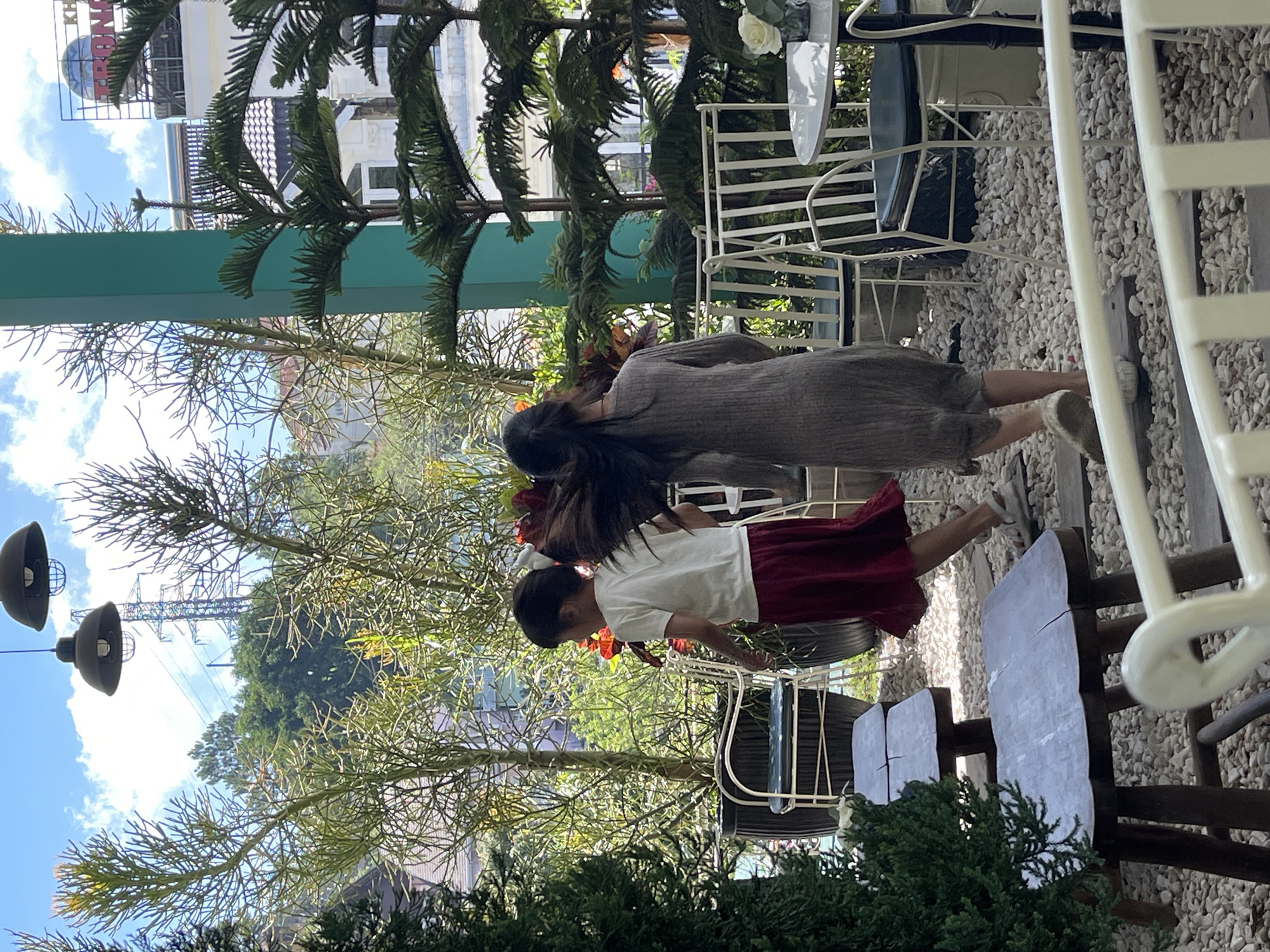 Two females are on a terrace, seemingly in the city of Da Lat, Vietnam. One female wears a gray dress, while the other is in a white top and red skirt. Tables and chairs are arranged on the terrace, which is covered with gravel. Green trees surround the terrace, and a building is visible in the background under a blue sky with clouds.