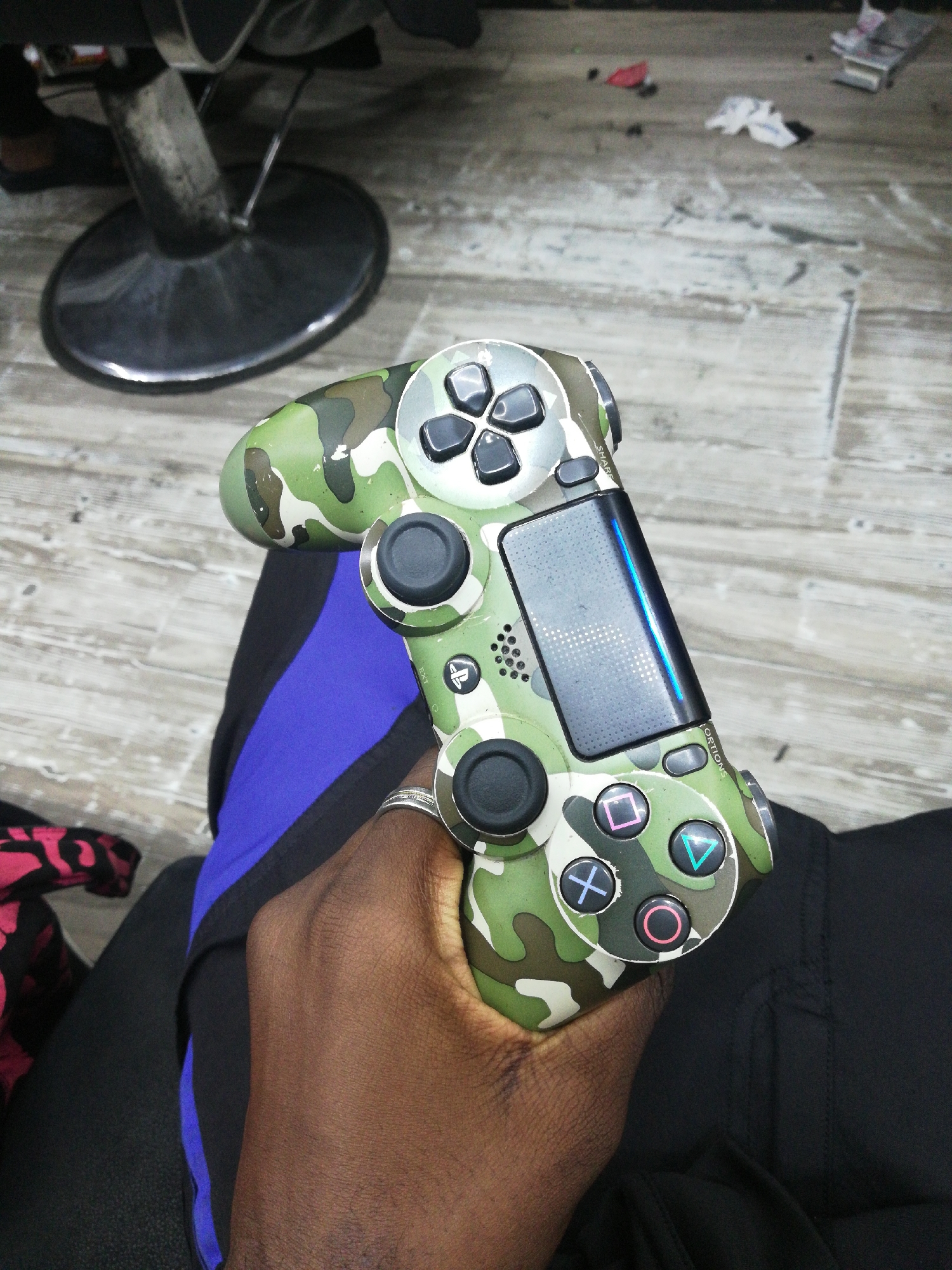 A person is holding a camouflage Playstation 4 controller. The person's hand is dark-skinned, and they are wearing a ring on their finger. The controller is green, brown, and white camouflage. The person is wearing black pants with a blue stripe on the side. The background is a tiled floor, with a barber chair and some hair clippings on the floor. The location is most likely a barbershop in Lagos, Nigeria.