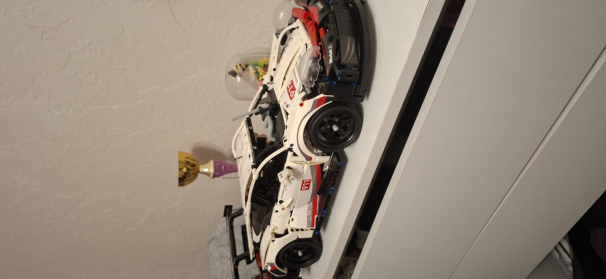 The image shows a white Lego Technic Porsche 911 model car on a white shelf in what appears to be a domestic setting. The car is detailed with red and black accents, racing decals including "911" and "PORSCHE", and the "Mobil 1" logo. A small trophy with a gold cup and a purple base is visible behind the car, along with a glass dome containing a decorative bee. The background is a textured, off-white wall. The bottom of the image shows the edge of a white cabinet. There are no people or any indication of the city of Shakhtarsk, Ukraine in the image.