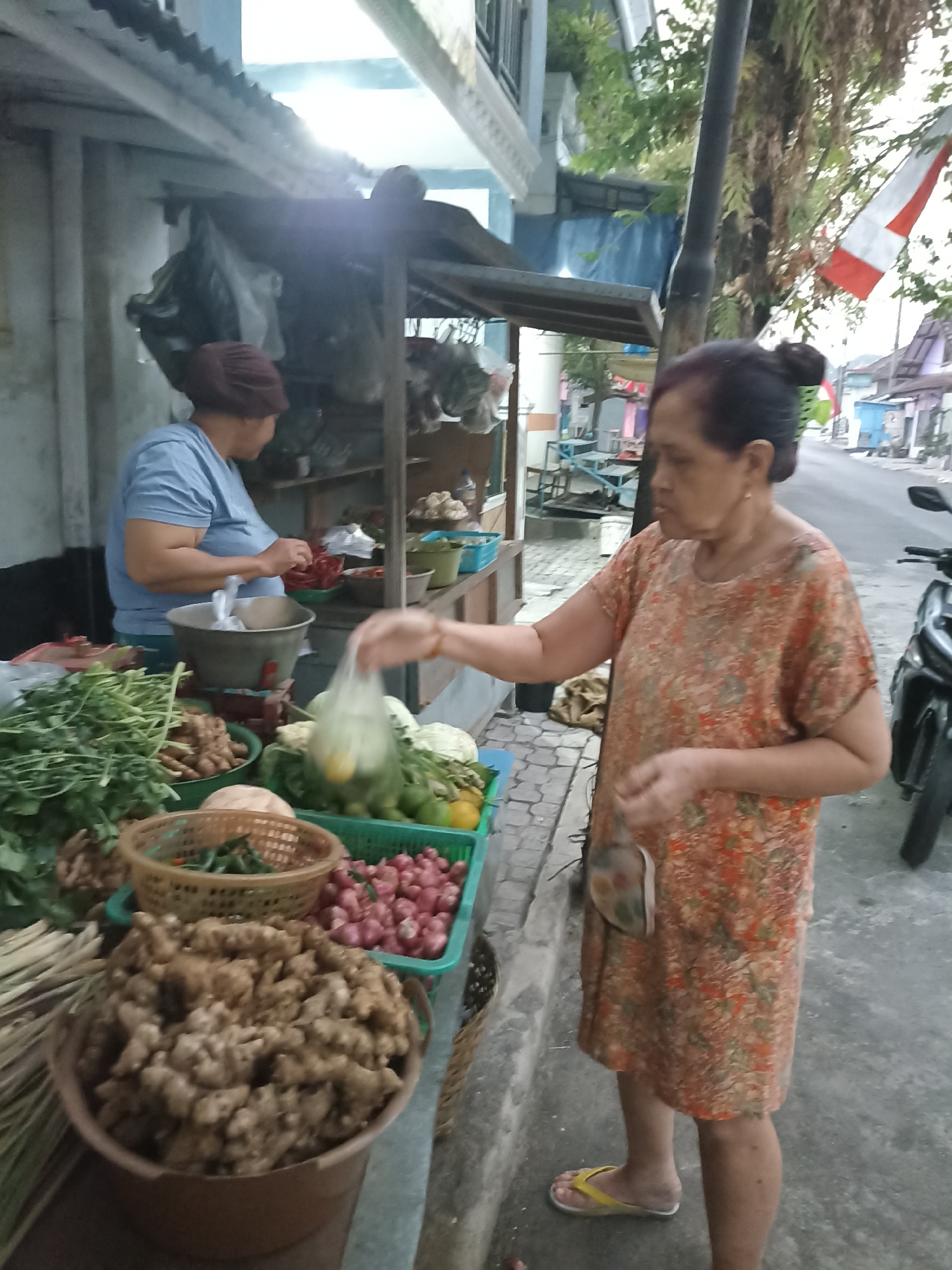 The image depicts an outdoor scene, likely a street market, in Lumajang, Indonesia. In the foreground, a woman in an orange floral dress is seen interacting with a vendor. She is holding a plastic bag and appears to be purchasing items from a stall. The stall is laden with various fresh produce, including large quantities of ginger, onions, and leafy greens. A second woman, wearing a blue t-shirt and a headscarf, is visible behind the stall, possibly the vendor. The background shows a street with a motorcycle parked on the right side, and a building with an Indonesian flag attached. The overall impression is a candid snapshot of daily life and commerce in a local Indonesian market.