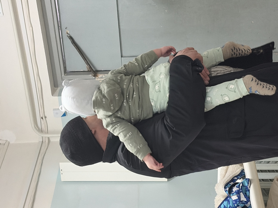 A person wearing a black beanie and a black jacket is holding a baby. The baby is wearing a white hat, a green top with white designs, green leggings, and beige socks with brown designs. The scene is indoors. A window is visible in the background. Laundry hangs nearby, suggesting a domestic setting. The photo is taken at an angle with the camera slightly off-center.