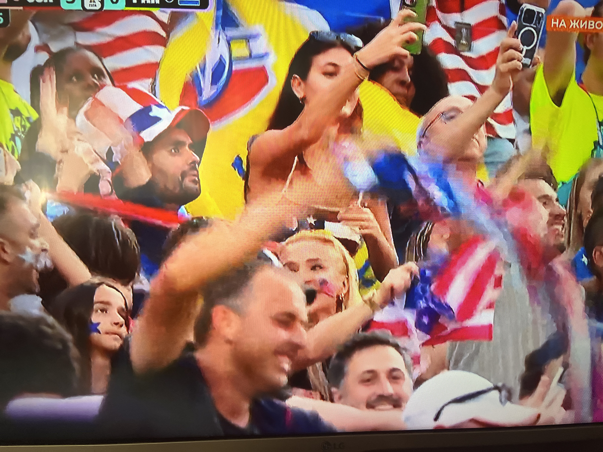 In an outdoor crowd, likely at a sporting event or festival, several people are visible, some with faces painted with American flags and stars. In the foreground, a man with a beard smiles, and to his right, another man with a mustache also smiles. Behind them, a young woman with long dark hair has a blue star painted on her cheek. Further back, a woman in a tan top is taking a selfie with her phone, while others around her hold up American flags. The atmosphere appears festive and energetic, with many people holding up phones to capture moments. The background is filled with blurred figures and colorful flags, predominantly red, white, and blue, suggesting a patriotic gathering. The lighting is bright, indicating daytime.