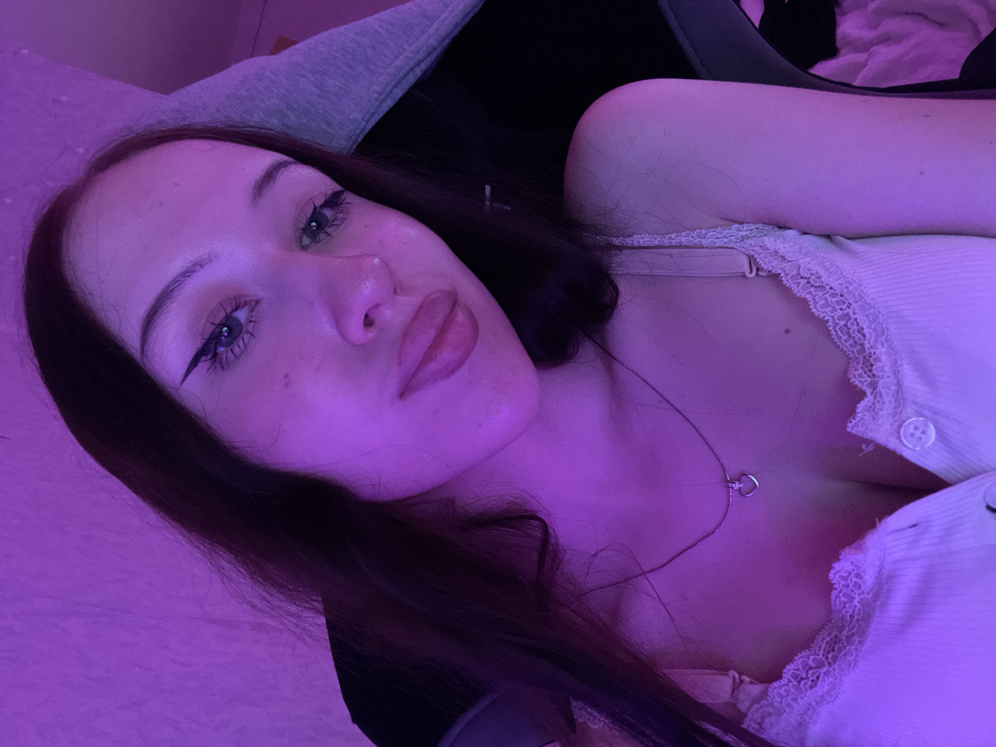The image is a close-up, high-angle selfie of a young woman illuminated by purple lighting. She has long, dark hair, dramatic winged eyeliner, and full lips. She is wearing a white tank top with lace trim and a delicate silver necklace with a heart pendant. The background is softly lit with purple, suggesting an indoor environment, possibly a bedroom or gaming setup. The image is likely taken in Votkinsk, Russia, based on the provided context, but no specific landmarks or elements within the image confirm this location.