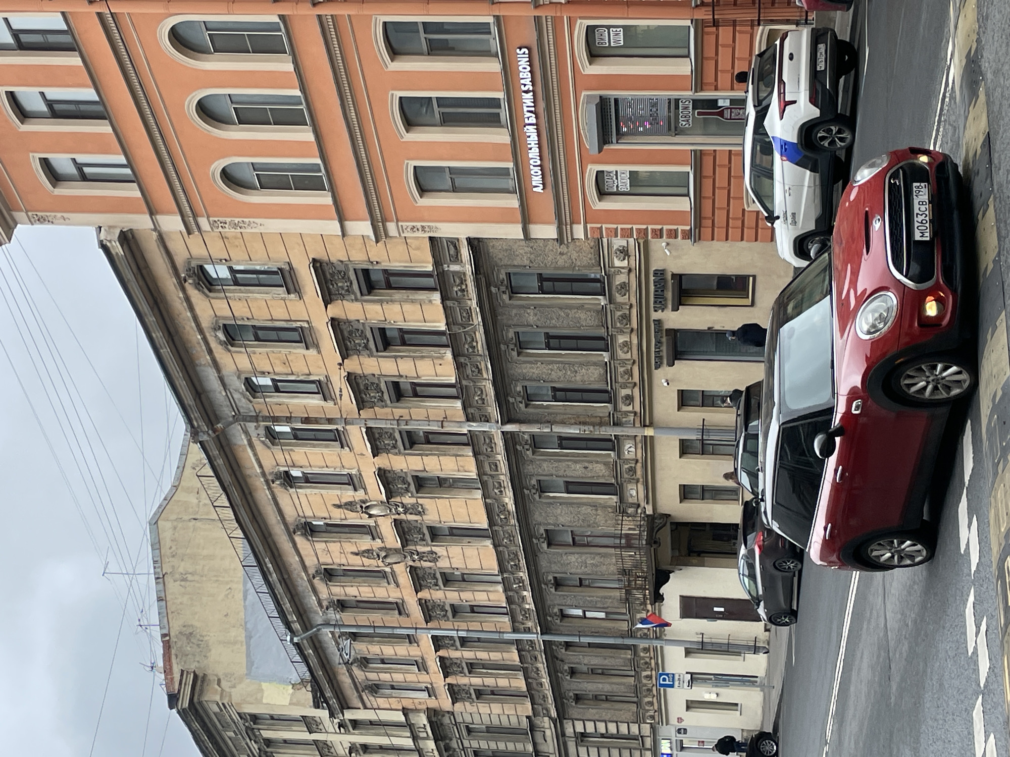 This image depicts a street scene in Saint Petersburg, Russia, featuring several buildings and parked cars. The buildings are characterized by their ornate facades and multiple rows of windows. The building on the right has a sign in Russian that translates to "Alcoholic Boutique Sabonis" and another that reads "Wine." A red Mini Cooper is parked on the right side of the street, with a partially visible license plate. Behind it is a white vehicle with a blue stripe. In the background, other cars are visible. The street is a typical urban setting with pedestrian walkways. The sky is overcast, indicating a cloudy day. There's a parking sign on the left side of the street and a Russian flag flying above a building entrance. People are walking along the sidewalks in the background.