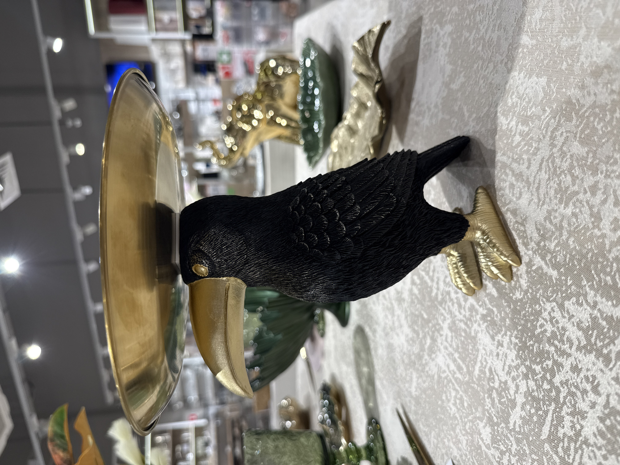 A close-up, eye-level shot focuses on a decorative black bird figurine with gold-colored feet and beak, balancing a gold-colored shallow dish on its head. The figurine is positioned on a textured, light-colored surface, possibly a tablecloth, with other decorative objects blurred in the background. These include green glass vases or bowls, a gold-colored elephant figurine, and a gold leaf-shaped dish. The background also shows shelving and lighting indicative of a retail environment. The scene is static, with no people or discernible actions. The image appears to be taken indoors, in a store. The location is not Izhevsk, Russia.