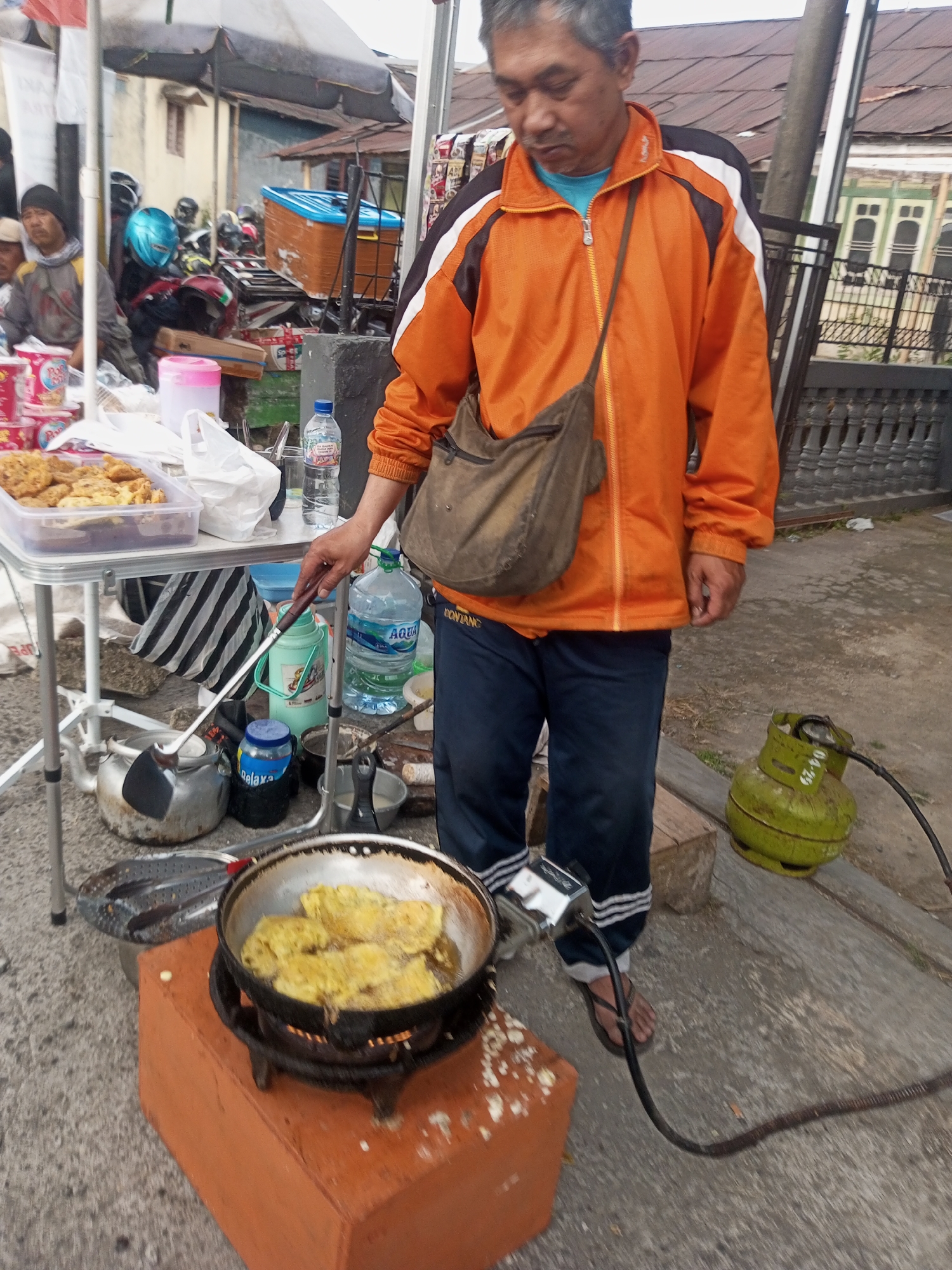 A man is cooking in Pandansari, Indonesia. He is wearing an orange jacket and blue pants, and is standing next to a portable stove where he is frying food in a pan. A gas cylinder is connected to the stove, and a hose runs from the cylinder to the stove. There are also several bottles of water and other items on a table next to the stove. The scene appears to be set up on the side of a road, with buildings and vehicles visible in the background. The ground is paved with concrete. The man is holding a spatula and is in the process of flipping the food in the pan.