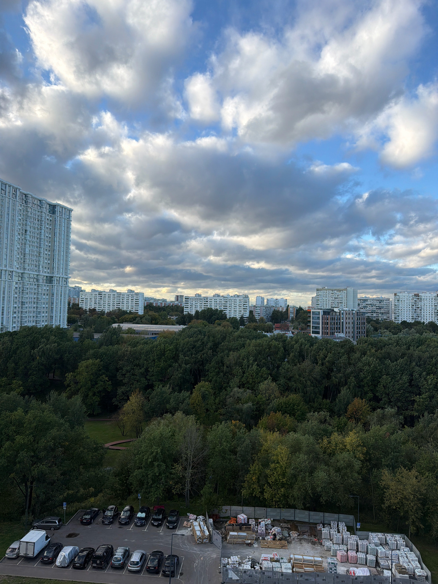 This is an eye-level, vertical, outdoor, daytime, medium shot of a cityscape with trees in Moscow, Russia, under a cloudy sky. In the foreground, a paved construction site is visible with stacks of building materials, including pallets of bagged goods, wooden planks, and rolls of material. Adjacent to the construction site is a parking lot with several cars and a white cargo van. A dense green forest dominates the middle ground, with a clearing containing a park-like area with a winding path. Beyond the trees, a series of residential buildings of various sizes and architectural styles are spread across the horizon, with one particularly tall, light-colored apartment building on the left. The sky is filled with a mix of dark and light clouds, with patches of blue sky peeking through.