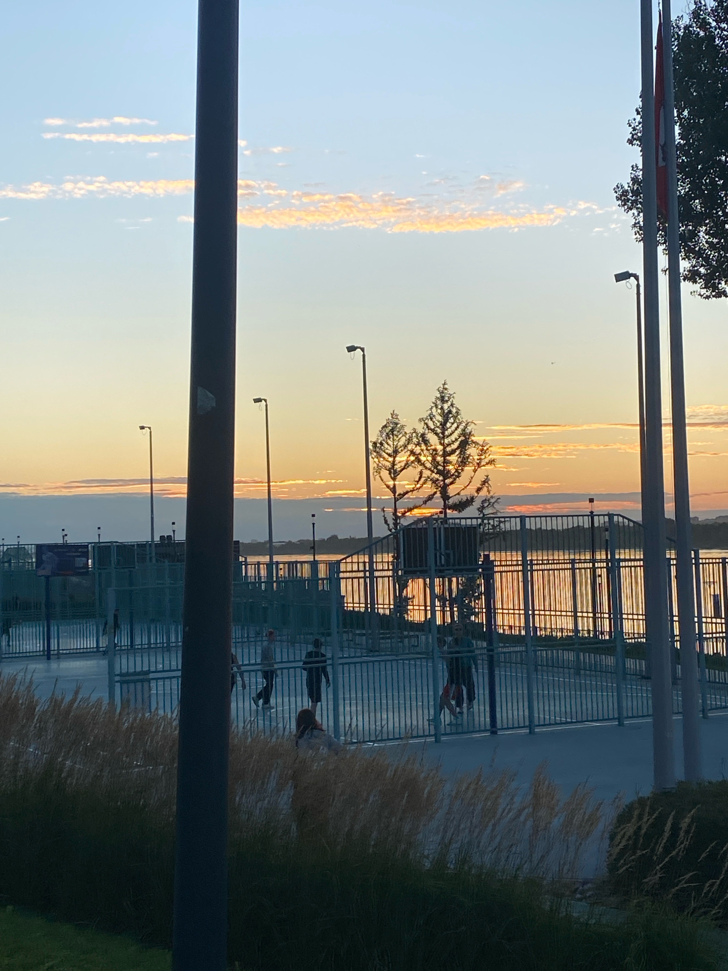 This image captures an outdoor scene at dusk, likely in the city of Perm, Russia, near a body of water. In the foreground, tall, dry grass and dark green bushes obscure the lower portion of the frame. Dominating the left side is a thick, dark pole.
The middle ground reveals a fenced sports court, possibly a basketball court, with a blue surface. Several people are visible within the court, some appearing to be walking or standing. One person sits in the lower right quadrant of the court area. Beyond the court is a metal fence bordering a wide body of water, reflecting the warm hues of the sunset. Two slender trees with distinctive crowns are silhouetted against the sky, with the setting sun partially visible behind them.
The background features a hazy skyline with a few buildings visible in the distance. The sky is a soft blue, transitioning to orange and pink clouds as it nears the horizon, indicating the end of daylight. Several tall lampposts are positioned around the sports facility, and two flags, one predominantly red, fly on poles to the right of the frame, with a portion of a dark green tree visible behind them. The overall atmosphere is tranquil and indicative of a recreational area at the close of day.