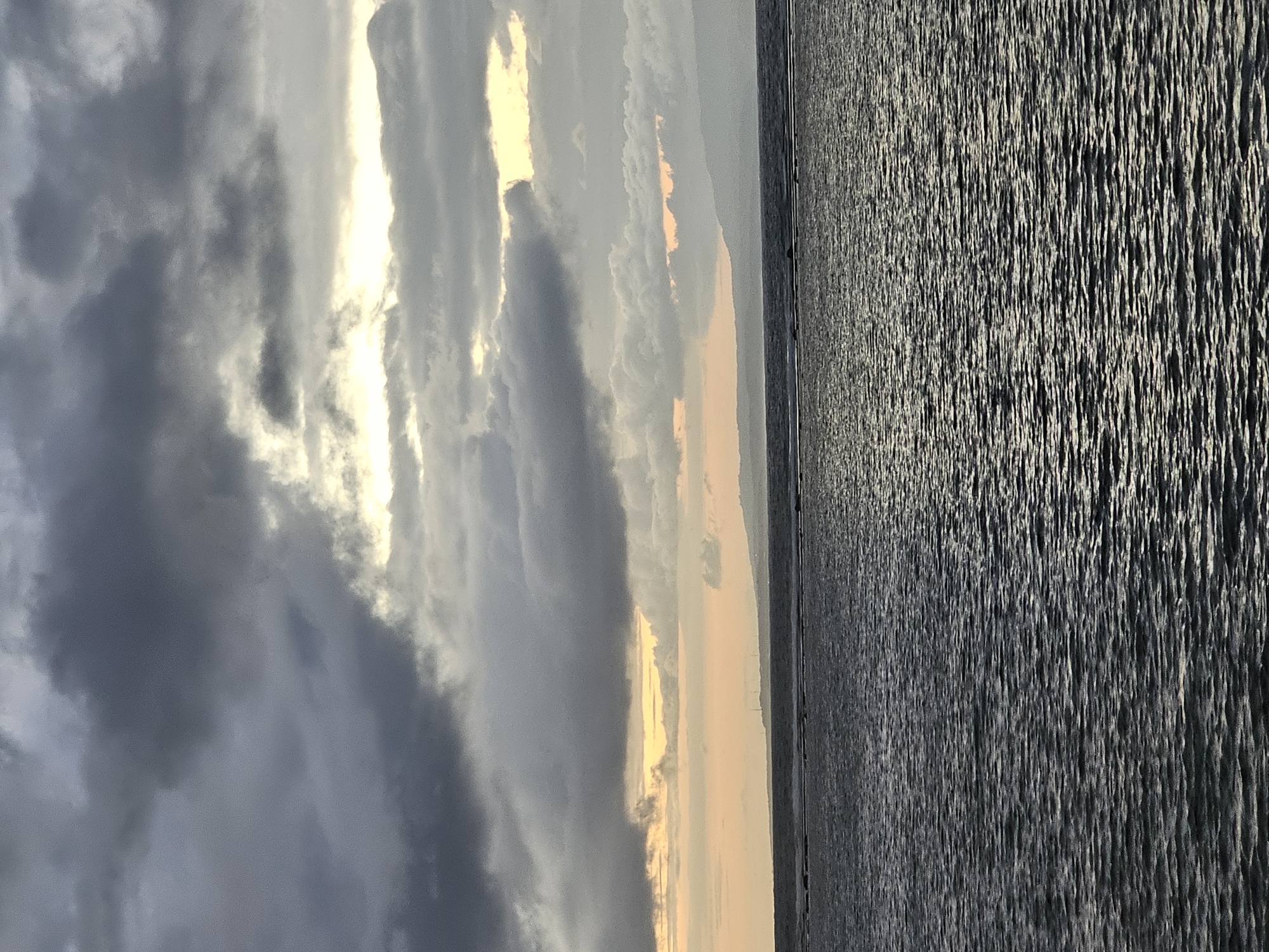 This image depicts a dramatic sky over a body of water. The sky is filled with a mix of dark, ominous clouds and brighter, more illuminated areas that suggest sunlight breaking through. Wispy, lighter clouds are interspersed with heavier, darker formations, creating a textured and dynamic atmosphere. A sliver of pale orange and yellow light is visible near the horizon, hinting at either sunrise or sunset.
The water occupies the right side of the frame and appears choppy, with the surface reflecting the light from the sky in numerous small, shimmering lines. The texture of the water suggests a gentle to moderate ripple. The horizon line is visible in the distance, separating the sky from the water, and appears relatively flat.
The scene evokes a sense of atmospheric tension and natural beauty. The contrast between the dark clouds and the illuminated sky, along with the textured water, creates a visually striking image. The time of day is likely early morning or late evening due to the light conditions. While specific location cues are not immediately apparent, the image captures a broad, open expanse of sky and water, common to coastal or lakeside environments. No people, objects, or visible text are present in the frame. The overall emotion conveyed is one of awe at the power and beauty of nature.