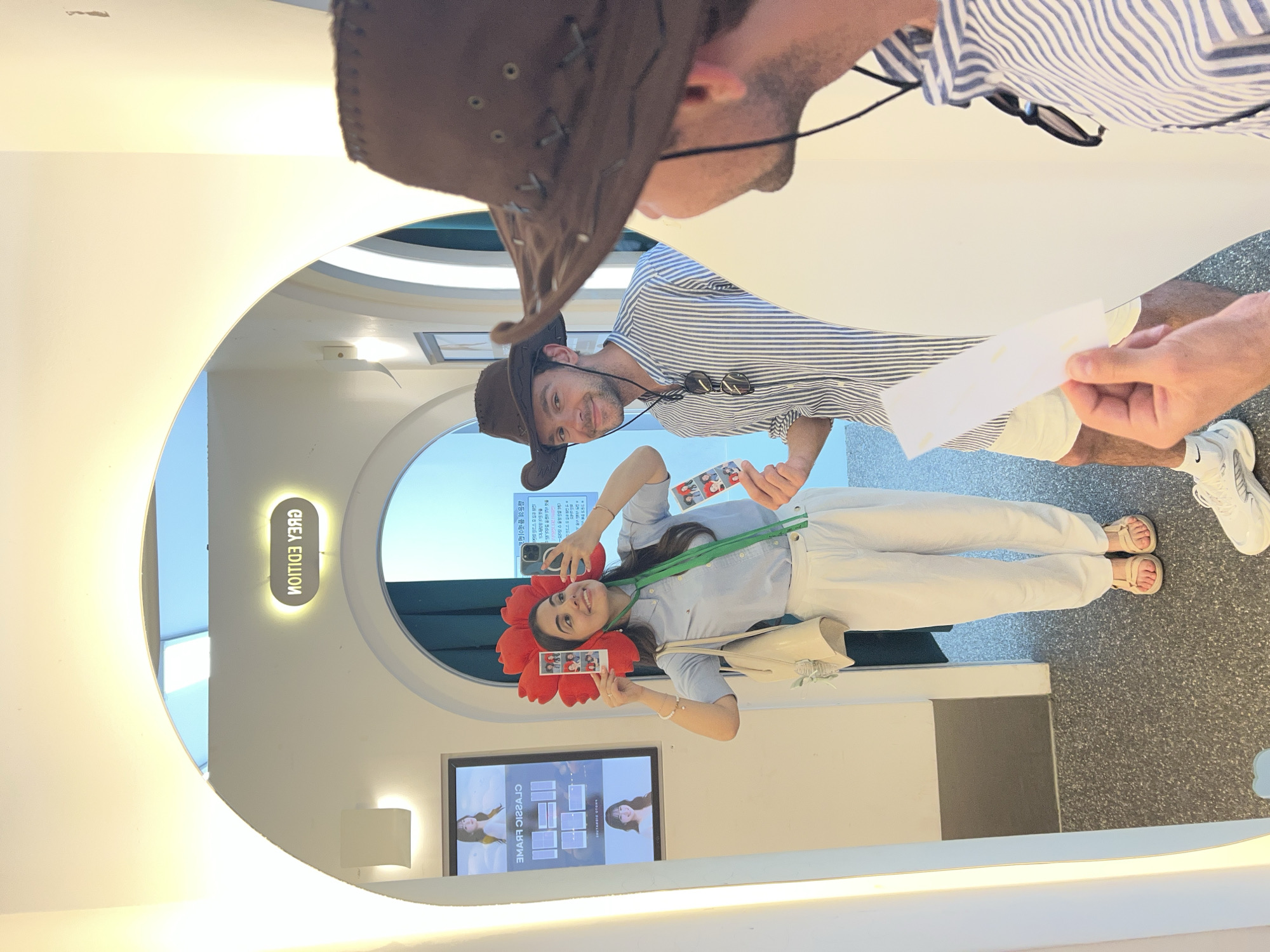 The image depicts a scene with a mirror reflecting a woman, a man, and the surrounding environment. The woman is wearing a large red flower headpiece and holding up a photo strip. She is dressed in a light blue shirt and white pants. The man in the reflection is wearing a brown cowboy hat and a striped shirt. He is holding a white piece of paper or ticket.
The setting appears to be a photo booth or studio, as indicated by the mirror setup and a screen displaying photo options. There's signage visible in the reflection, including the text "GREA EDITION" in vertical letters. A TV screen at the bottom left displays photos.
The picture was most likely taken in the city of Seoul, South Korea, as the text displayed on the screen is in Korean.