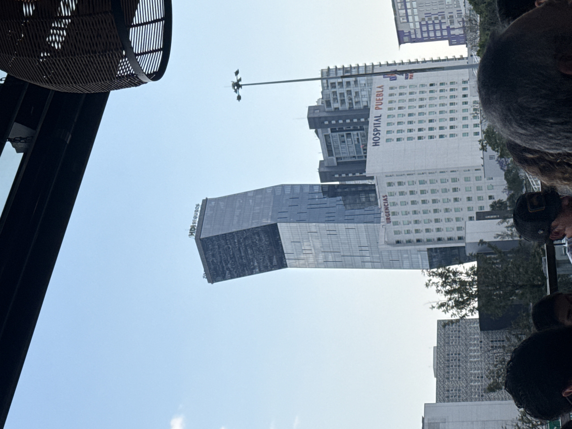 The image shows a view looking up at several buildings in an urban environment. Visible structures include a modern skyscraper with a dark, blocky section, a white building labeled "HOSPITAL PUEBLA", and other tall buildings in the background. A tall light post extends from the hospital building. In the foreground, out-of-focus objects suggest an indoor setting, possibly a restaurant or cafe. People are partially visible on the right side of the frame. The location appears to be in Puebla, given the signage on the hospital building.