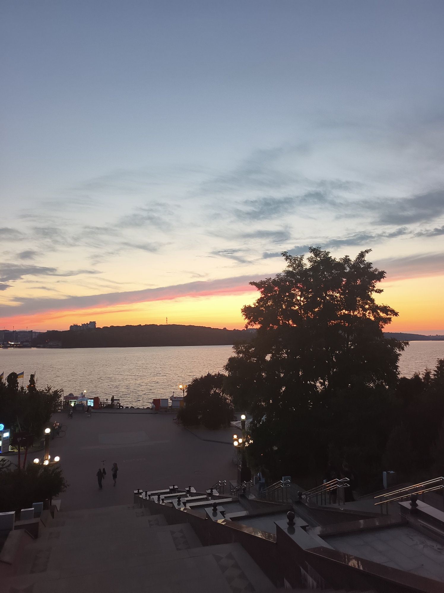 The image depicts a panoramic view of the Ternopil Pond during sunset. The sky is painted with hues of orange, pink, and purple, with scattered clouds. A silhouette of a large tree dominates the right side of the frame, with its branches extending across the sunset. In the foreground, a wide staircase with decorative railings leads down towards a promenade along the waterfront. Several people are visible on the promenade, some walking and others standing. Buildings and structures are visible on the opposite bank of the pond, and a few lights are lit along the waterfront, indicating the evening hour. The overall atmosphere is serene and picturesque, capturing the beauty of a sunset over the city.