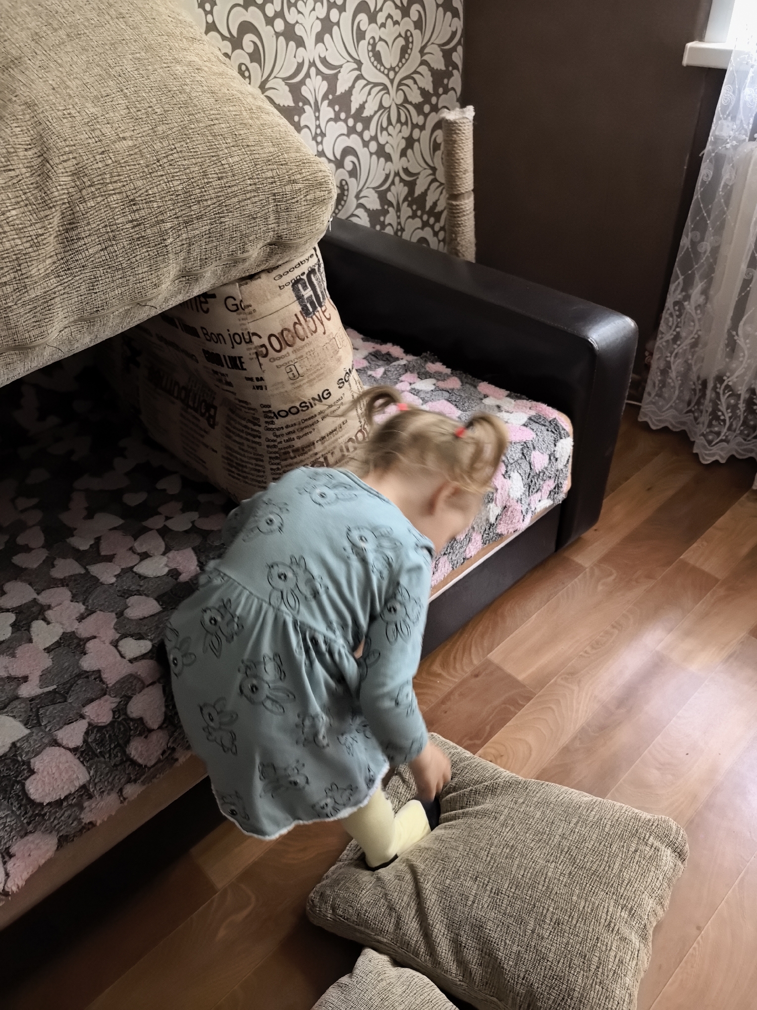 An indoor scene shows a young child in a blue dress with a rabbit pattern, standing on a wooden floor next to a couch. The couch has dark brown leather arms and is covered with a blanket featuring a pattern of pink and gray hearts. A large, light brown textured cushion is placed on the floor in front of the couch, and another similar cushion is propped up against the couch. The child is bent over, looking down towards the cushion on the floor, with their blonde hair in two pigtails. Behind the couch, there is a wall covered in patterned wallpaper with large floral designs and a beige scratching post. A sheer white curtain is visible on the right side of the image.