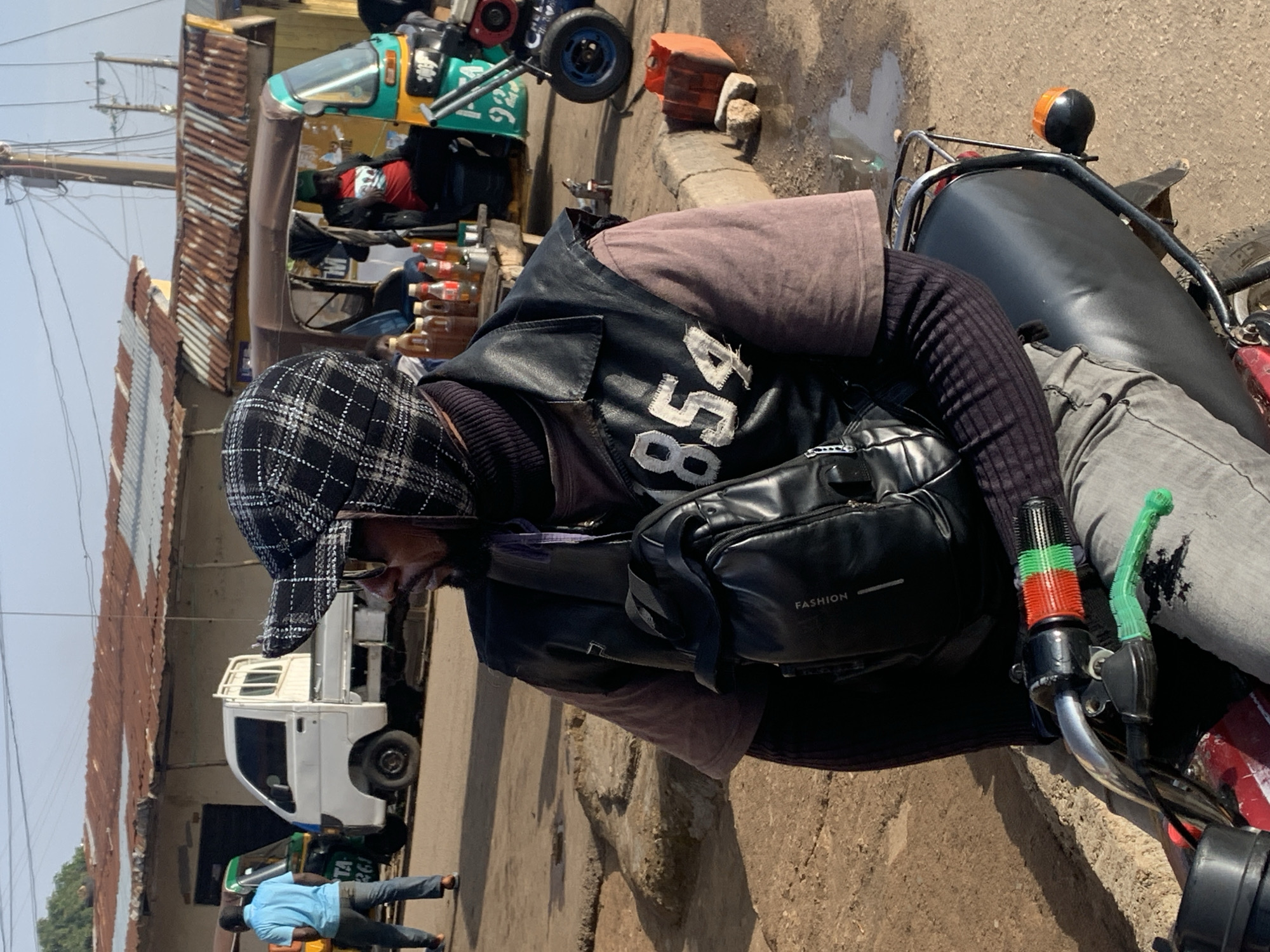 This image captures a scene in Gura Top, Nigeria, depicting a person resting on a motorcycle. The subject is wearing a black and white checkered cap, a dark vest with "854" printed in white on the back, a purple t-shirt, and grey pants. A black bag with the word "FASHION" visible is slung across the chest. The motorcycle appears to be a common model found in the region, possibly used for transport or as a business vehicle.
In the background, other vehicles are present, including a white truck and a colorful auto-rickshaw, suggesting a busy street or market area. The setting is outdoors, with a dirt road and some corrugated metal structures visible, indicating a potentially dusty environment. The lighting suggests it is daytime, likely mid-morning or afternoon, with bright sunlight casting shadows. The person on the motorcycle appears relaxed, possibly taking a break from work or travel. There are no explicit cues about emotions beyond a sense of stillness and repose. The overall impression is a snapshot of daily life in a vibrant Nigerian locale.