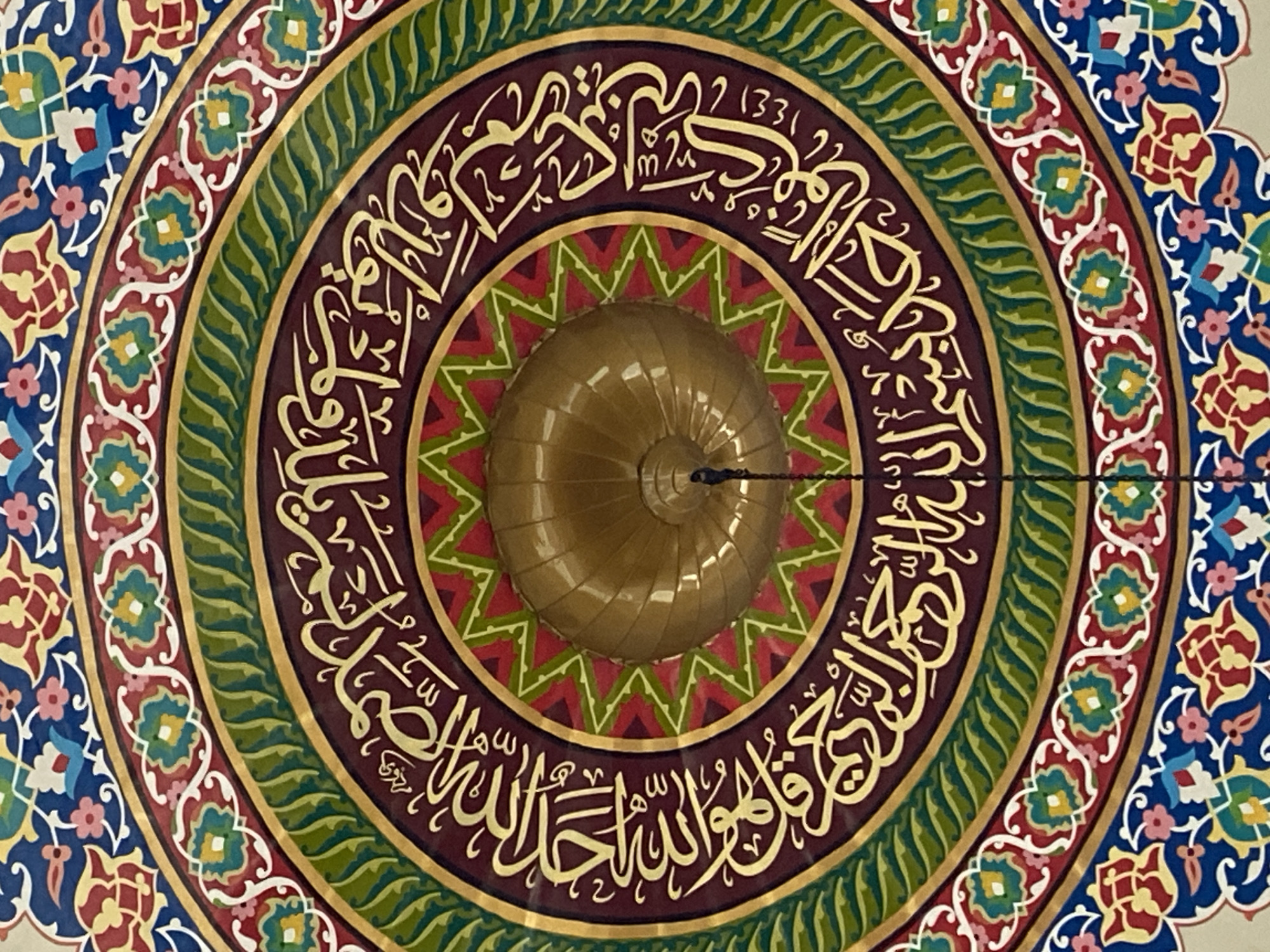 The image shows a detailed view of a circular ceiling decoration in a building. The design features concentric rings of intricate patterns and Arabic calligraphy in gold lettering. The outermost ring has a blue background with red, yellow, and green floral motifs. Inside this, there are bands of red, green, and gold patterns, with the calligraphy predominantly featured on a deep red band. At the center of the ceiling is a golden dome. The overall impression is one of ornate Islamic artistry. The image does not provide any information about its location in Lhokseumawe, Indonesia.