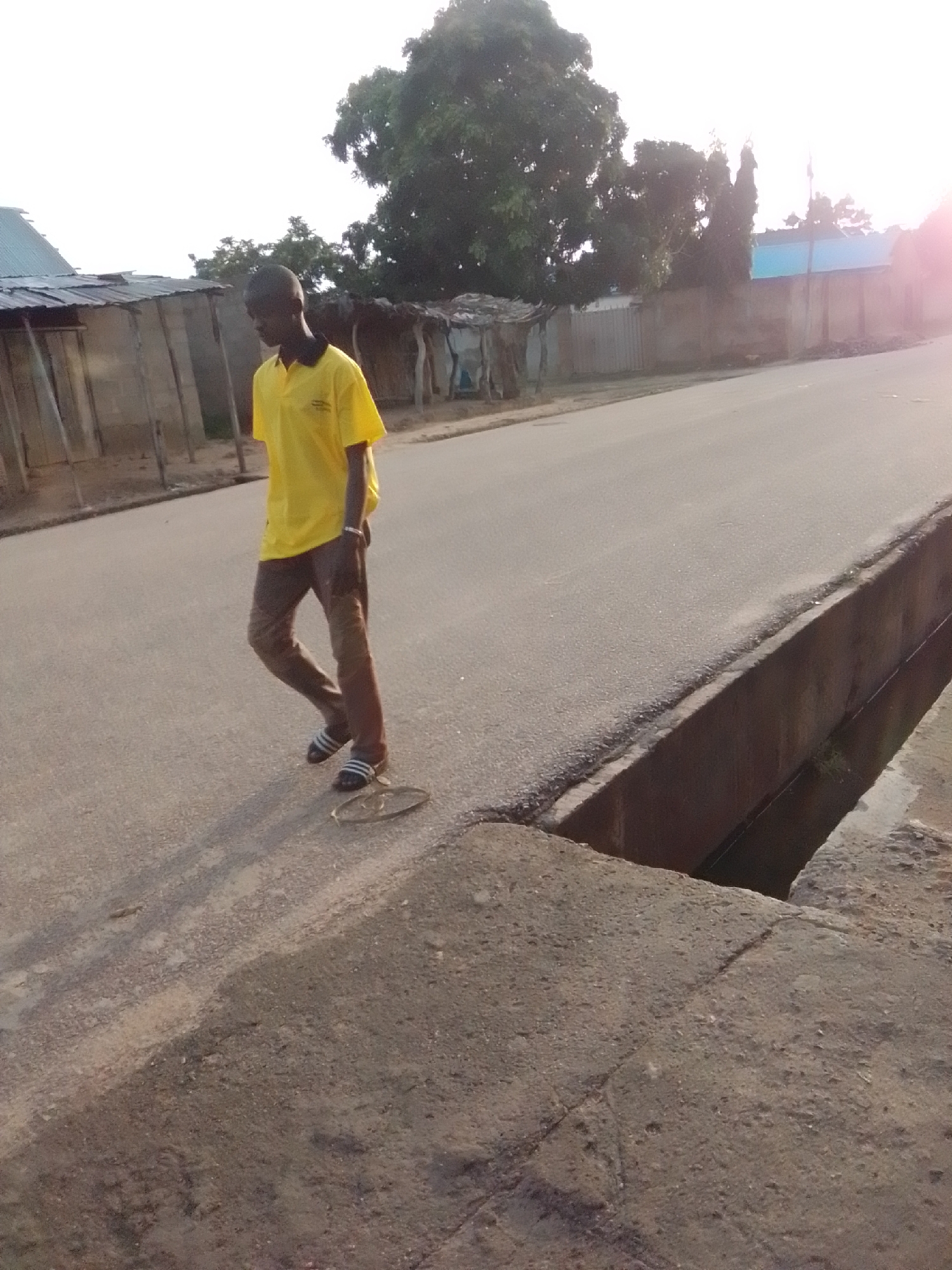 A young Black male wearing a yellow polo shirt and brown pants walks along a paved road. He is wearing black and white striped sandals. A circular object with a woven texture is on the road near his feet. The road runs alongside a concrete drainage ditch. In the background, there are several structures with corrugated metal roofs and walls made of wood and other materials. Trees and vegetation are visible behind the structures. The sky is bright, suggesting it is daytime. The scene appears to be in a rural or semi-urban area, possibly in the city of Gombe, Nigeria, given the architectural style of the buildings and the presence of the drainage ditch.
