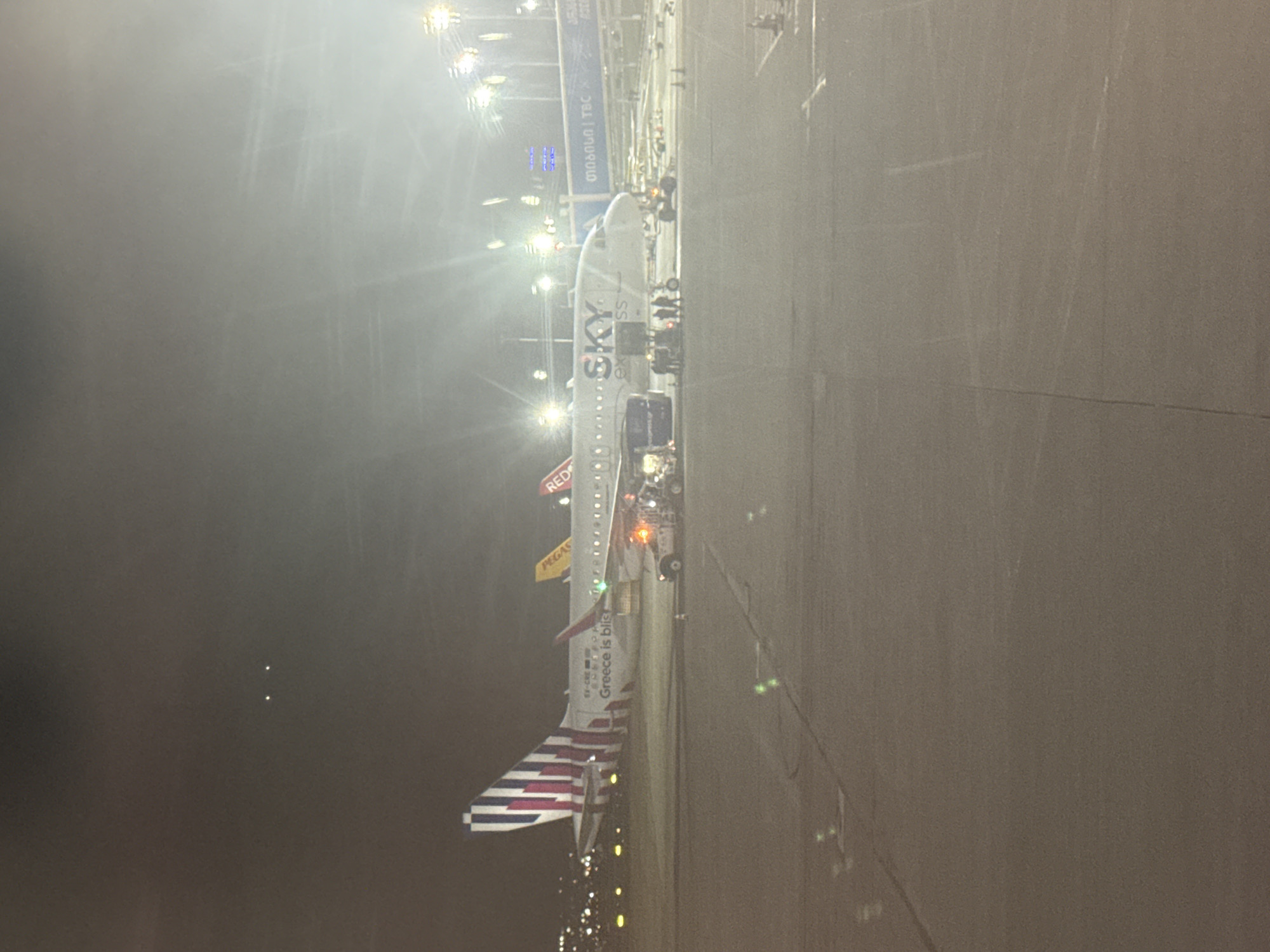 The image shows an Airbus A320 aircraft parked at Tbilisi International Airport (TBS) in Tbilisi, Georgia. The aircraft is painted in the livery of Sky Express, a Greek airline. Ground personnel and vehicles are visible around the aircraft. The photo was taken at night or during low-light conditions, as indicated by the artificial lighting and foggy or hazy atmosphere. The terminal building is visible in the background, with signage in Georgian and English.