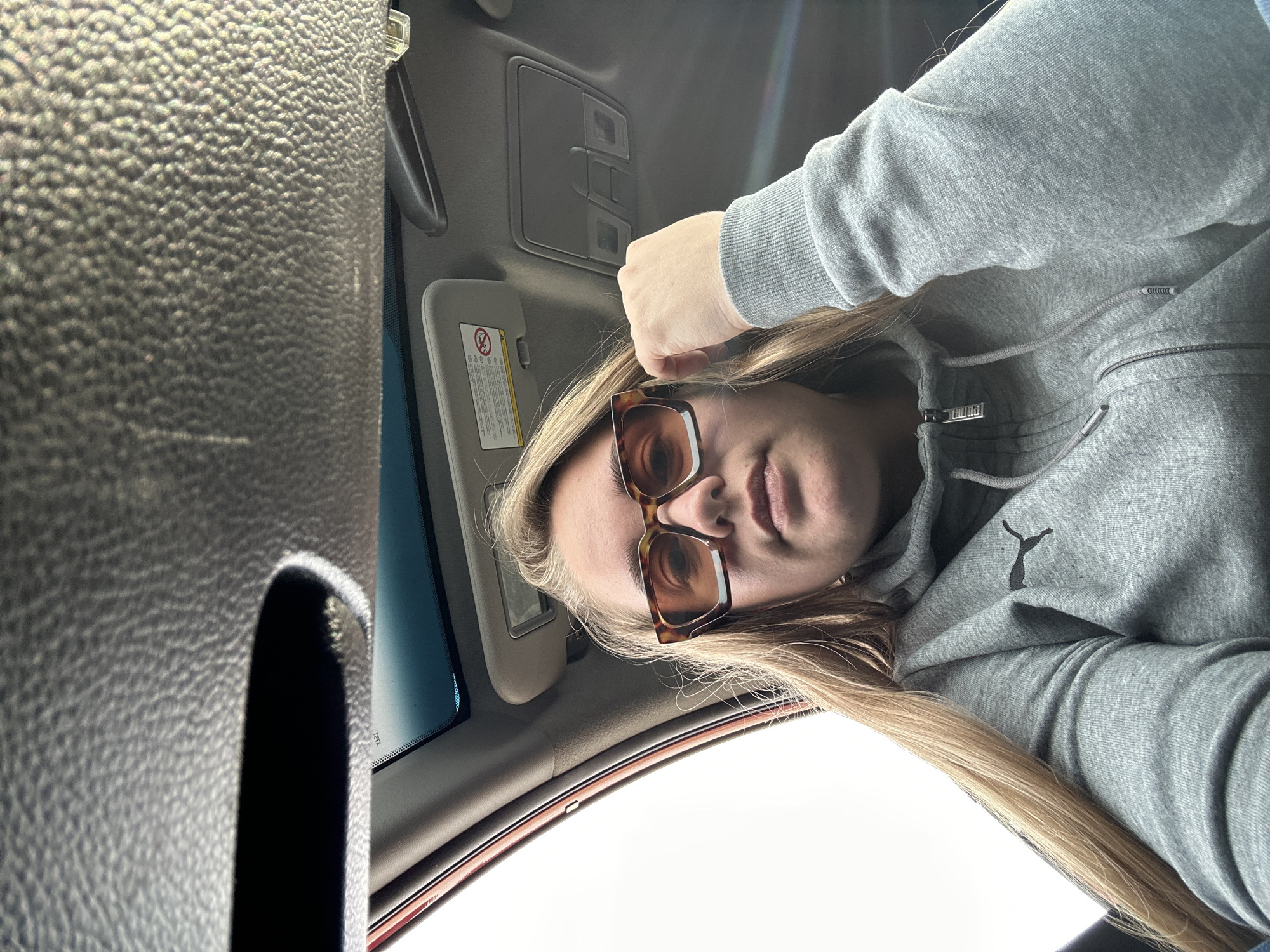 A female is positioned inside a vehicle. She is wearing a grey Puma hoodie and brown tortoise-shell eyeglasses. Her blonde hair is visible. Her hand is resting on the interior roof of the car. The car's sun visor is down. The car appears to be in Ural'skoe, Russia based on the request. The specific location in the city is not discernible.