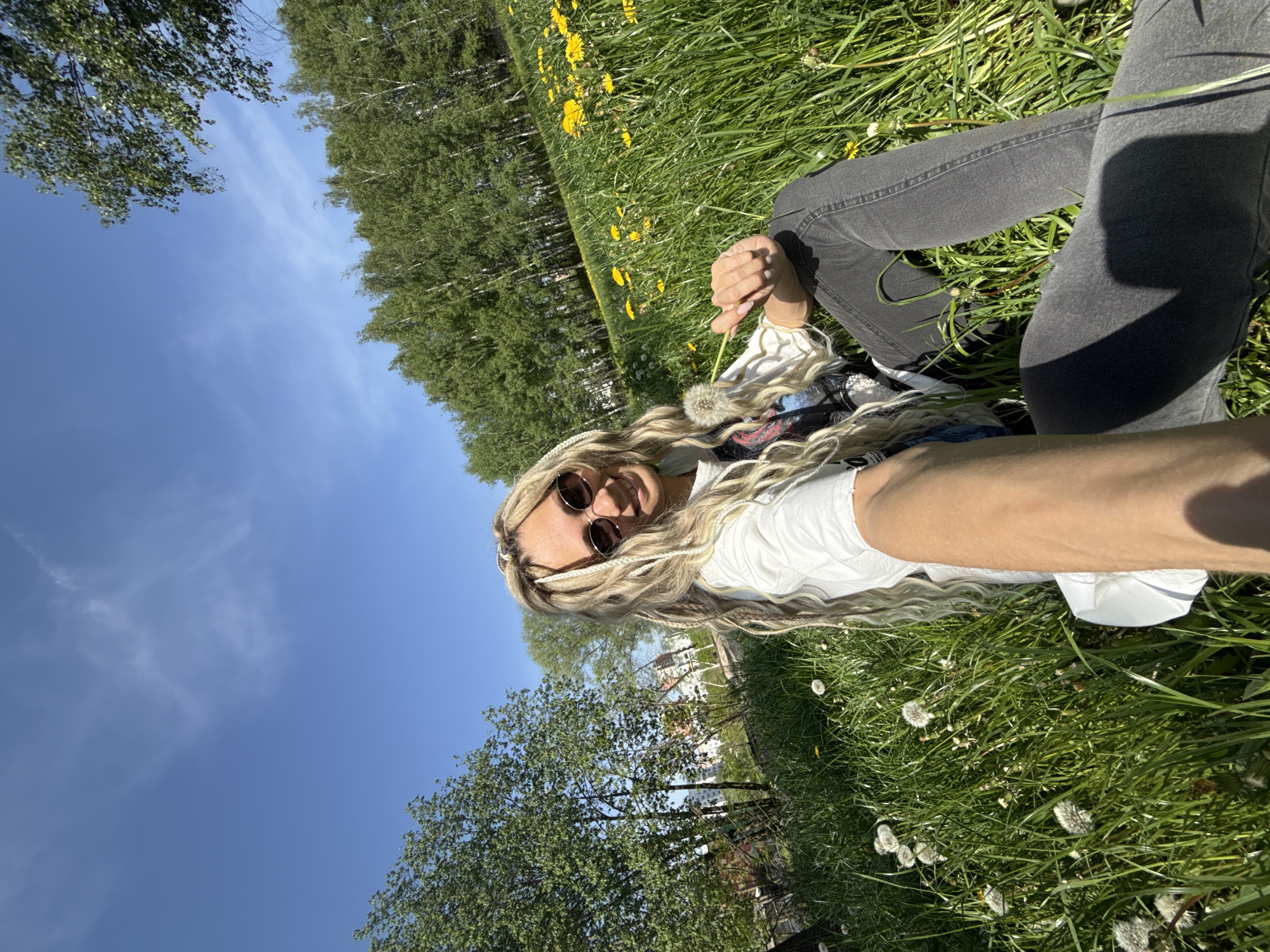 A person with blonde hair, wearing sunglasses, a white shirt, and gray jeans is sitting in a grassy field. The field is filled with dandelions, both yellow and white seed heads. The person holds a dandelion seed head in their hand. Trees are visible in the background, along with a blue sky with some clouds. The photo appears to be taken from a low angle, possibly as a selfie. The exact location within the city of Заволжье, Russia is not discernible from the image.