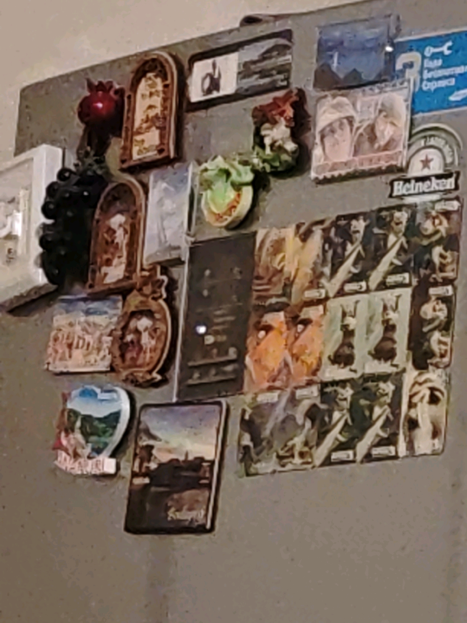 A collection of magnets adorns a metallic surface, possibly a refrigerator. The magnets depict various images, including religious icons, landscapes, and what appear to be portraits. One magnet, in the lower right quadrant of the image, shows a stylized representation of the city of Ninotsminda, Georgia, with a prominent church building and surrounding landscape. The overall impression is one of collected mementos or souvenirs. The lighting suggests an indoor setting, and the background is a neutral, textured surface.