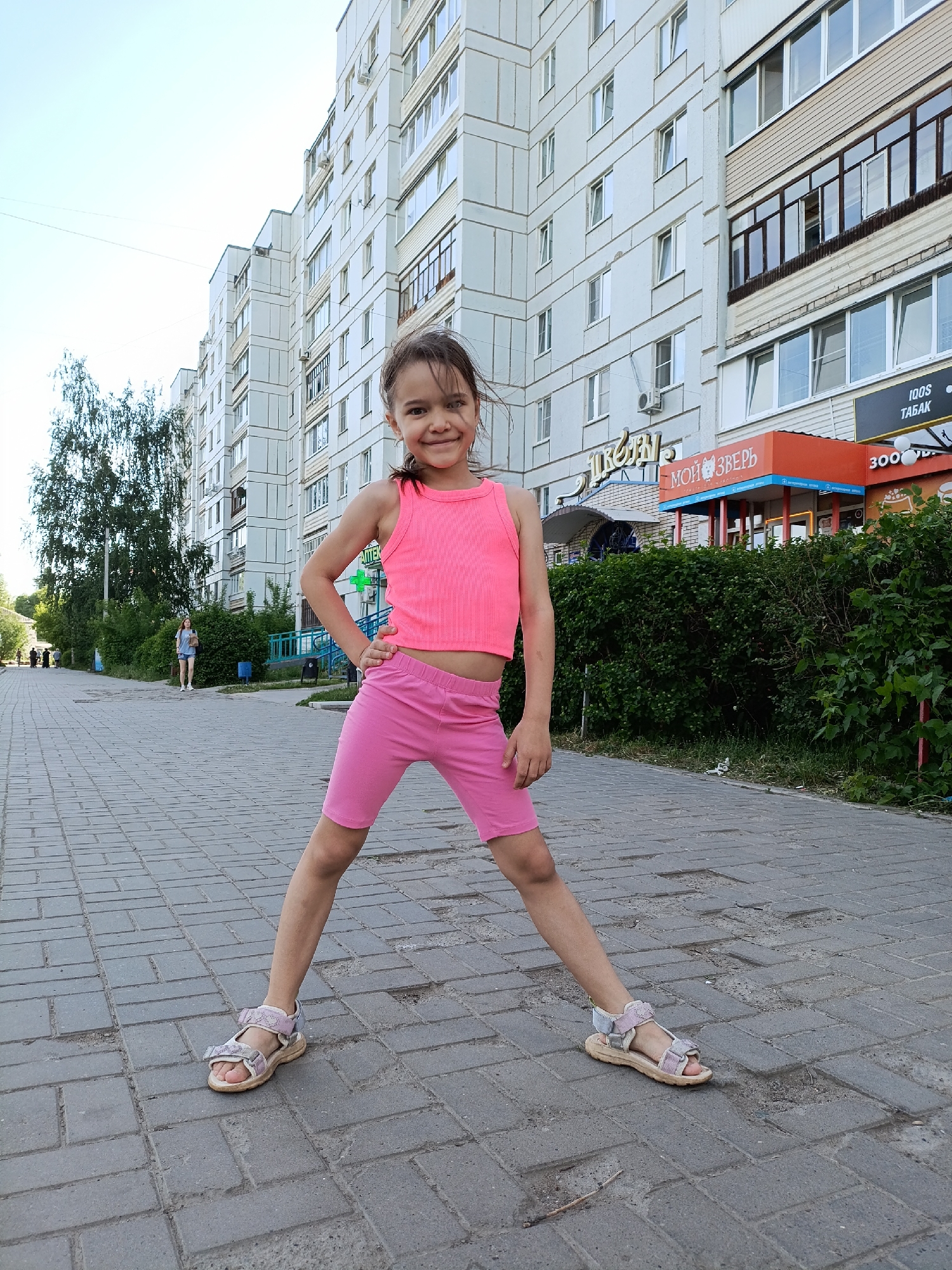 A young girl stands on a brick sidewalk in Chaikovsky, Russia, wearing a pink tank top, pink shorts, and sandals. She is posing with her legs wide apart and her left hand on her hip. Behind her is a multi-story apartment building with businesses at street level, including a store with a sign reading "Мой Зверь" (My Beast) and "IQOS Табак" (IQOS Tobacco). A green hedge is visible beside the sidewalk, and in the distance, a woman is walking away on the sidewalk. Other pedestrians are walking in the distance. The sky is bright and clear.