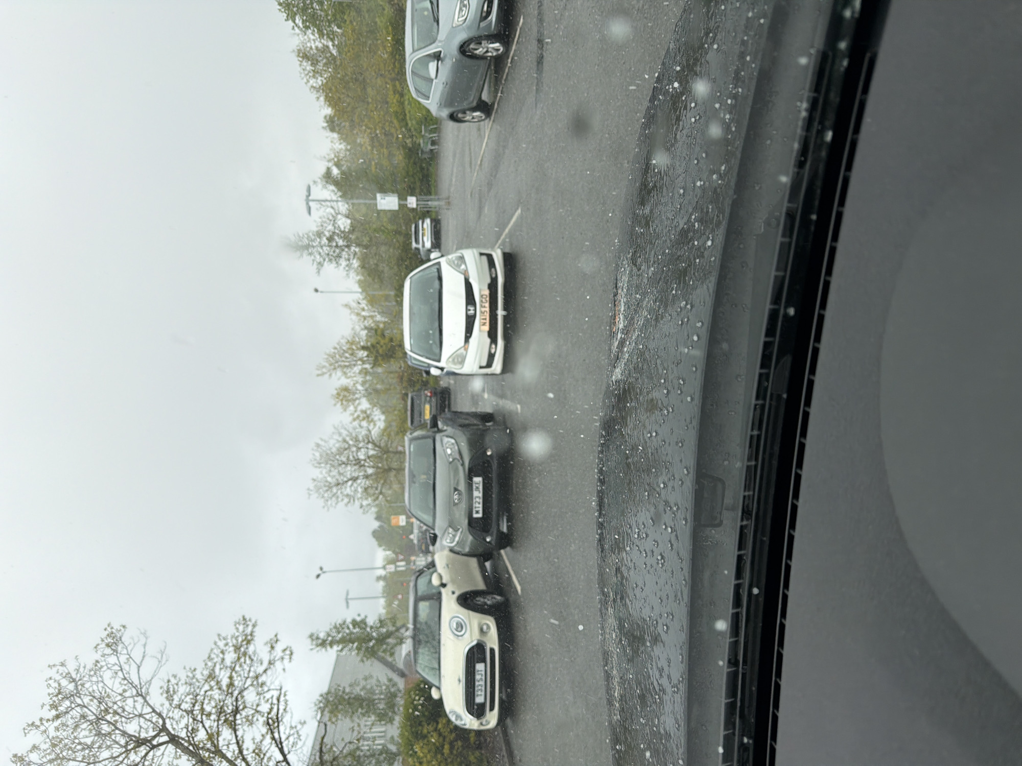 The image shows a view from inside a vehicle, possibly parked, at a car park in Pity Me, United Kingdom. The sky is overcast, and rain or hail is visible on the windshield. Several cars are parked in a row in the parking spaces. License plates visible include T33 SJT, KT23 JKL and N115 FGD. Trees and light poles are visible in the background. There is no evidence of people or activity other than parked cars.