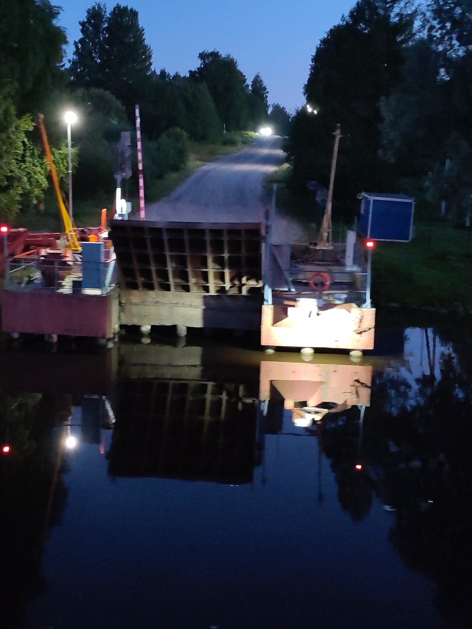 The image depicts a nighttime scene at Annensky Most, Russia, featuring a ferry crossing. The ferry, a flat-bottomed vessel, is partially illuminated, with lights visible on its deck and on surrounding structures. A road leads away from the ferry landing, with a railroad crossing signal visible on the right side of the road. Trees and vegetation line the sides of the road and the waterway, reflecting in the dark water. The sky is dark, indicating it is nighttime. The ferry appears to be equipped with a ramp or bridge that is raised, suggesting it is not currently in operation. A small blue structure, possibly a control booth or storage unit, is situated on the ferry's deck. The overall impression is one of a functional but quiet transportation hub at night.