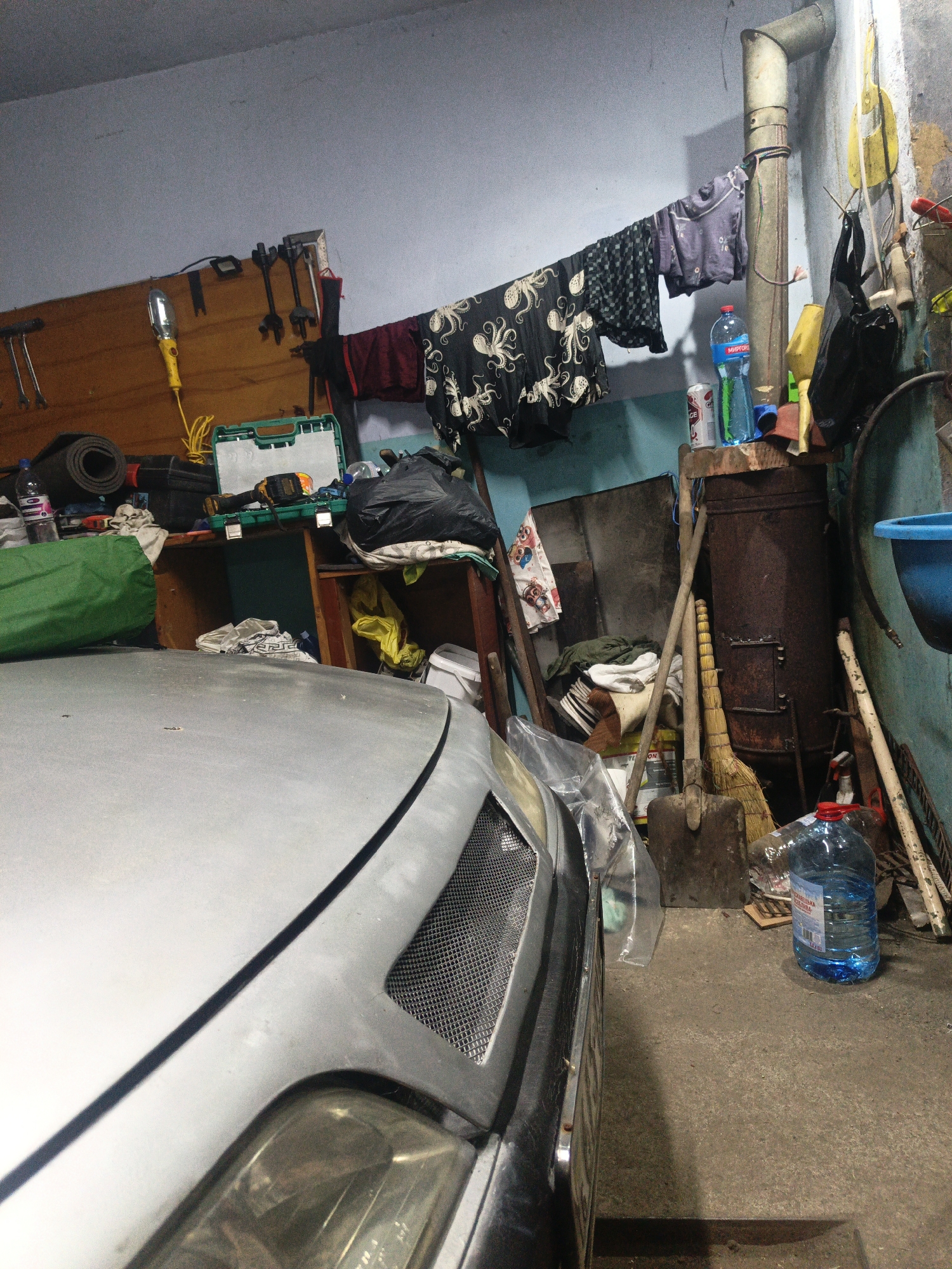 The image depicts a cluttered garage in Sychavka, Ukraine. A silver car occupies the foreground, with its hood partially visible. The garage interior is filled with various items, including tools hanging on a wooden board, a green tool case, black trash bags, clothing draped on a clothesline, a rusty metal barrel, a broom, and shovels. Several plastic bottles, including a large blue one, are also present. The walls are painted a light blue, and the floor is concrete. The overall impression is one of disarray and storage.