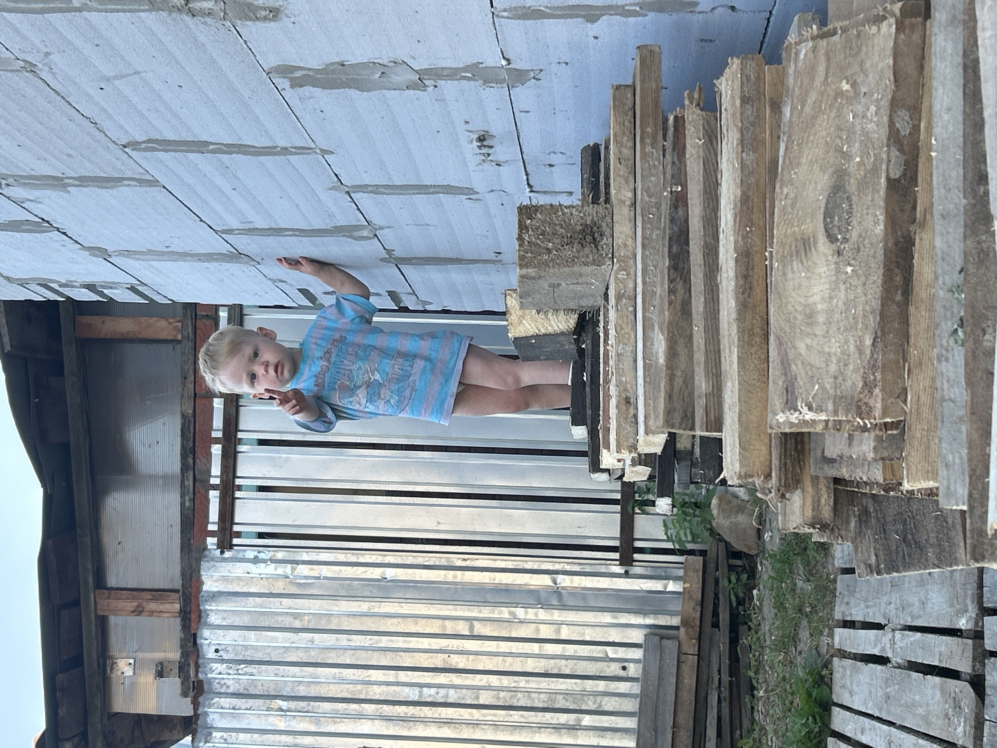 The image shows a young Caucasian boy with blond hair standing on a pile of lumber in front of a light blue cinder block wall. The boy is wearing a striped t-shirt and shorts, and he is holding up two fingers on his right hand. To the left of the boy is a corrugated metal fence and a wooden structure. The ground is covered in grass and more lumber. The scene appears to be outdoors, possibly in a backyard or construction area. The sky is visible in the upper portion of the image. This image was taken in Chaikovsky, Russia.