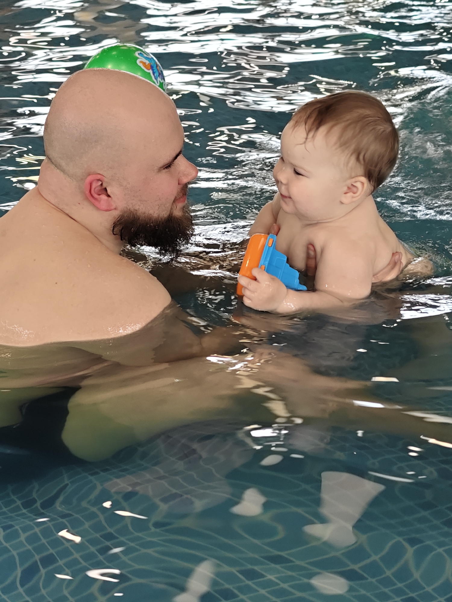 This image depicts a heartwarming interaction between a man and a baby in an indoor swimming pool. The man, with a bald head and a dark beard, is submerged chest-deep in the water, looking directly at the baby with a gentle smile. The baby, with light brown hair, is positioned in front of the man, supported by him, and holds a bright blue and orange plastic toy boat in their hands. Both subjects are smiling and making eye contact, conveying a sense of joy, affection, and engagement in their activity.
The setting is clearly an indoor swimming pool, with the water showing distinct ripples and reflections on its surface. Through the clear water, the blue and white checkered tile pattern of the pool floor is visible, especially in the lower half of the image. A green and white inflatable ball, decorated with a colorful butterfly design, floats on the water behind the man's head. The scene suggests a relaxed and happy moment, likely a swimming lesson or playful session between a parent or guardian and an infant. No legible text is visible within the frame. The event takes place in what appears to be a well-lit facility, in Votkinsk, Russia.
