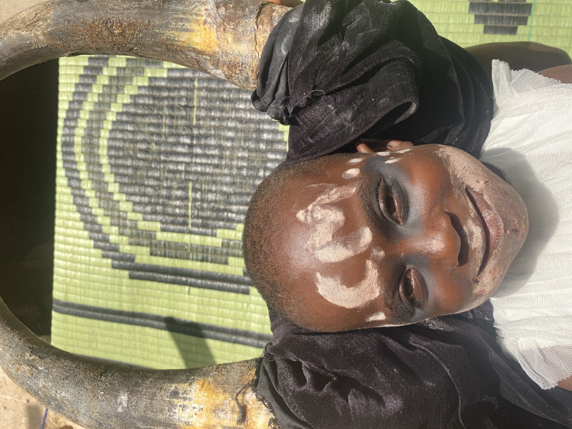 The image shows a close-up of a dark-skinned person lying on a woven mat inside a large, circular, wooden structure or frame. The person has closed eyes and appears to be decorated with white paint on their face. They are draped in both black velvet-like fabric and white tulle. The location is Ufu Esioni, Nigeria.