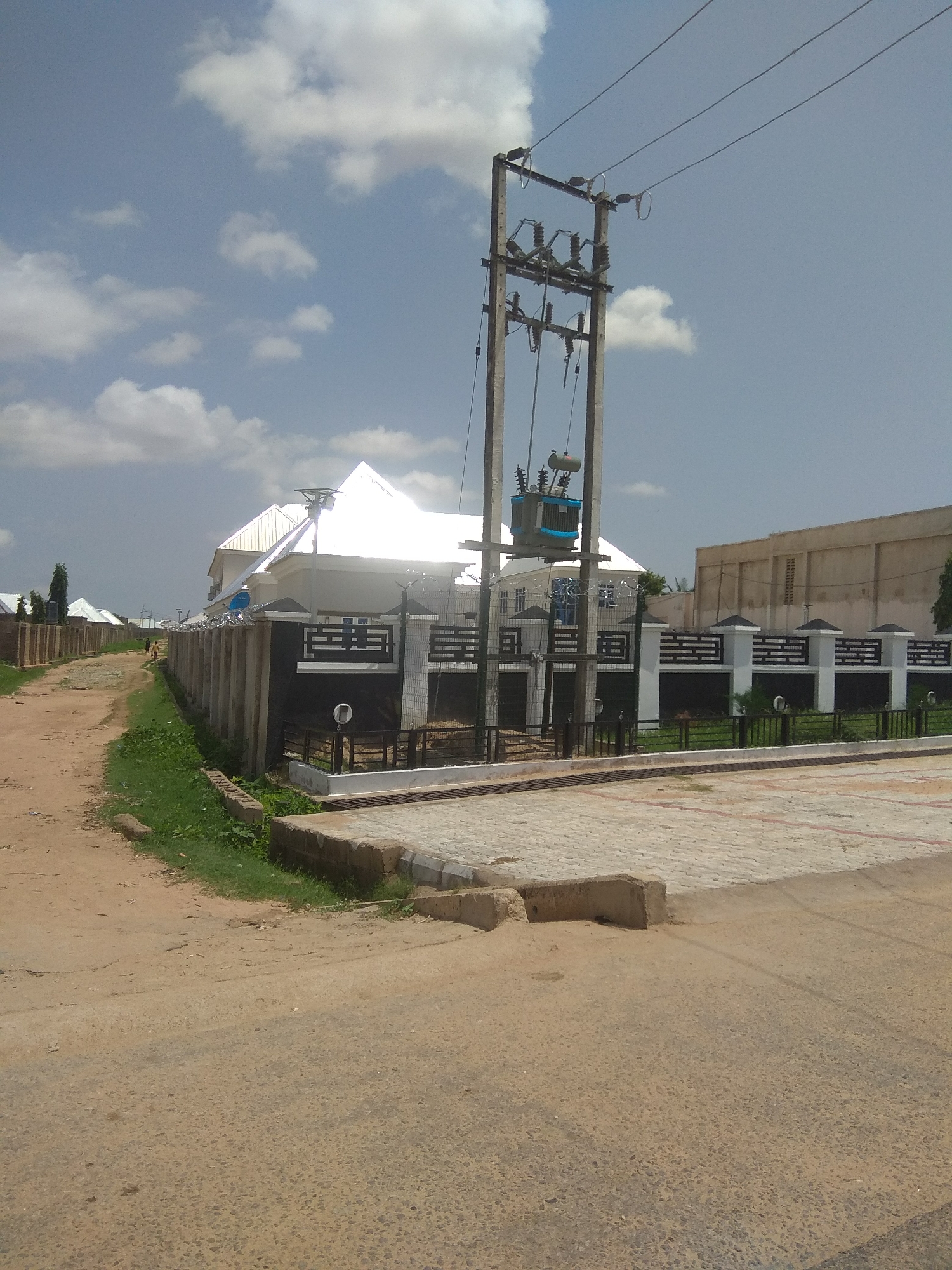 The image depicts a street scene in Jalingo, Nigeria, under a partly cloudy sky. In the foreground, a dirt road with some patchy vegetation lines the right side. To the right of the dirt road, a paved area with interlocking bricks is visible. Further into the scene, a modern building with a white, pitched roof and dark decorative accents on its facade is enclosed by a concrete and black metal fence topped with barbed wire. Adjacent to this property, a power pole with a transformer and electrical wires stands prominently. Beyond the main building, other structures and fences are visible along the street. Two individuals are walking on the dirt road in the distance. The environment appears to be a residential or developing urban area.