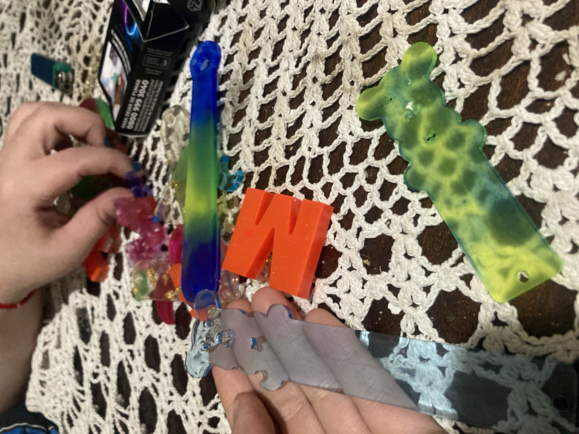 The image shows a collection of objects arranged on a lace doily, possibly on a table. These objects include:
* A lighter.
* A box with text that includes a phone number and the phrase "PARA DEJAR DE FUMAR".
* Several colorful resin items of various shapes, including a tall blue-to-green item, a giraffe, a clear item, and an orange block with the letter "N".
* Two hands are visible, one holding a clear item and the other interacting with the colorful resin items.
Given the phrase "PARA DEJAR DE FUMAR" on the box, it is likely located in a Spanish-speaking country. Considering that the location is specified as Buenos Aires, Argentina, it may be a depiction of an activity or an arrangement of items on a surface, potentially inside a home or business in Buenos Aires. The composition and variety of objects suggest a possible crafting or artistic activity.